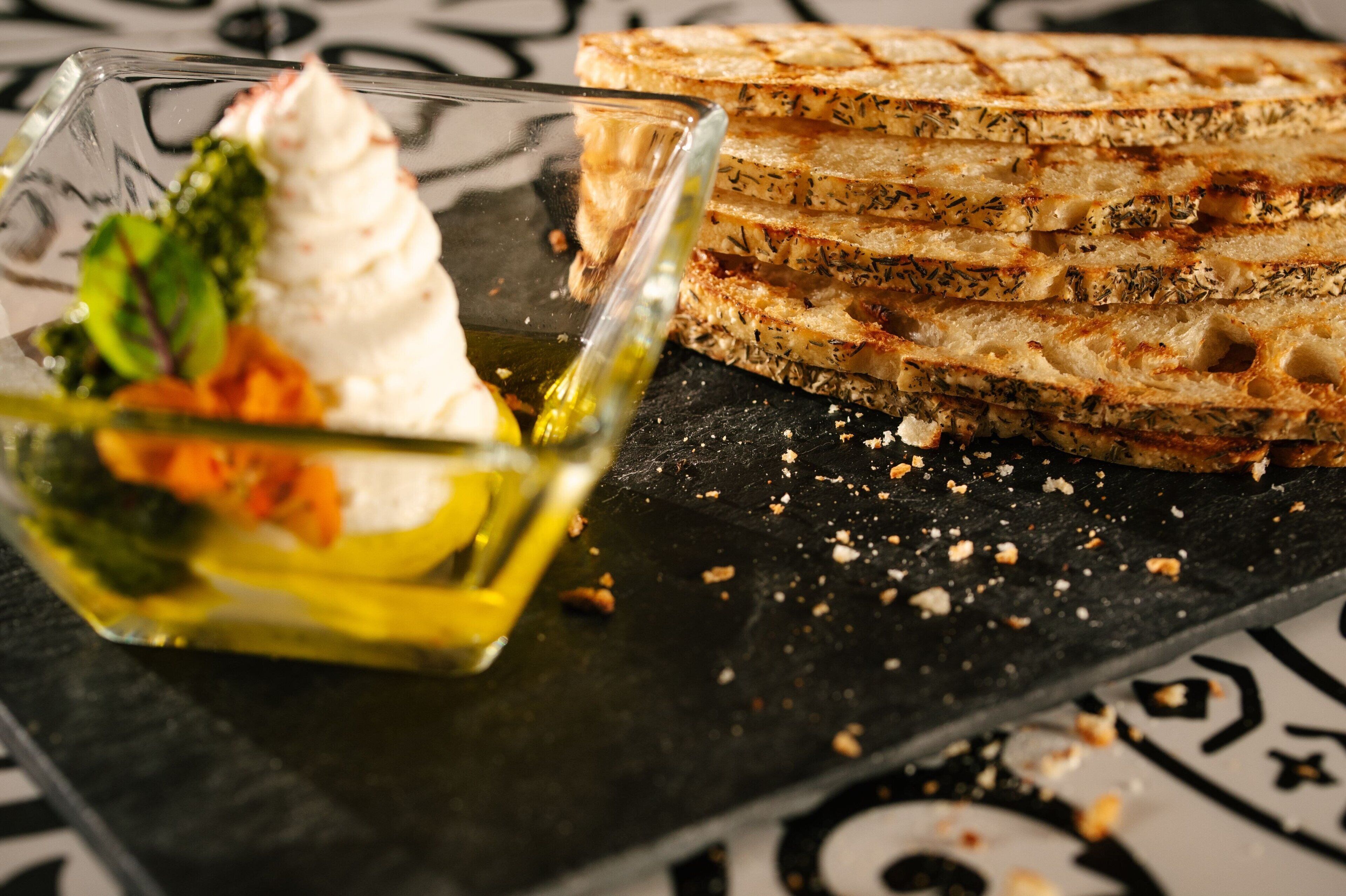 Una pila de pan tostado con hierbas, un vaso de aceite de oliva y un cuenco pequeño de ensalada con hojas verdes y rodajas de naranja.