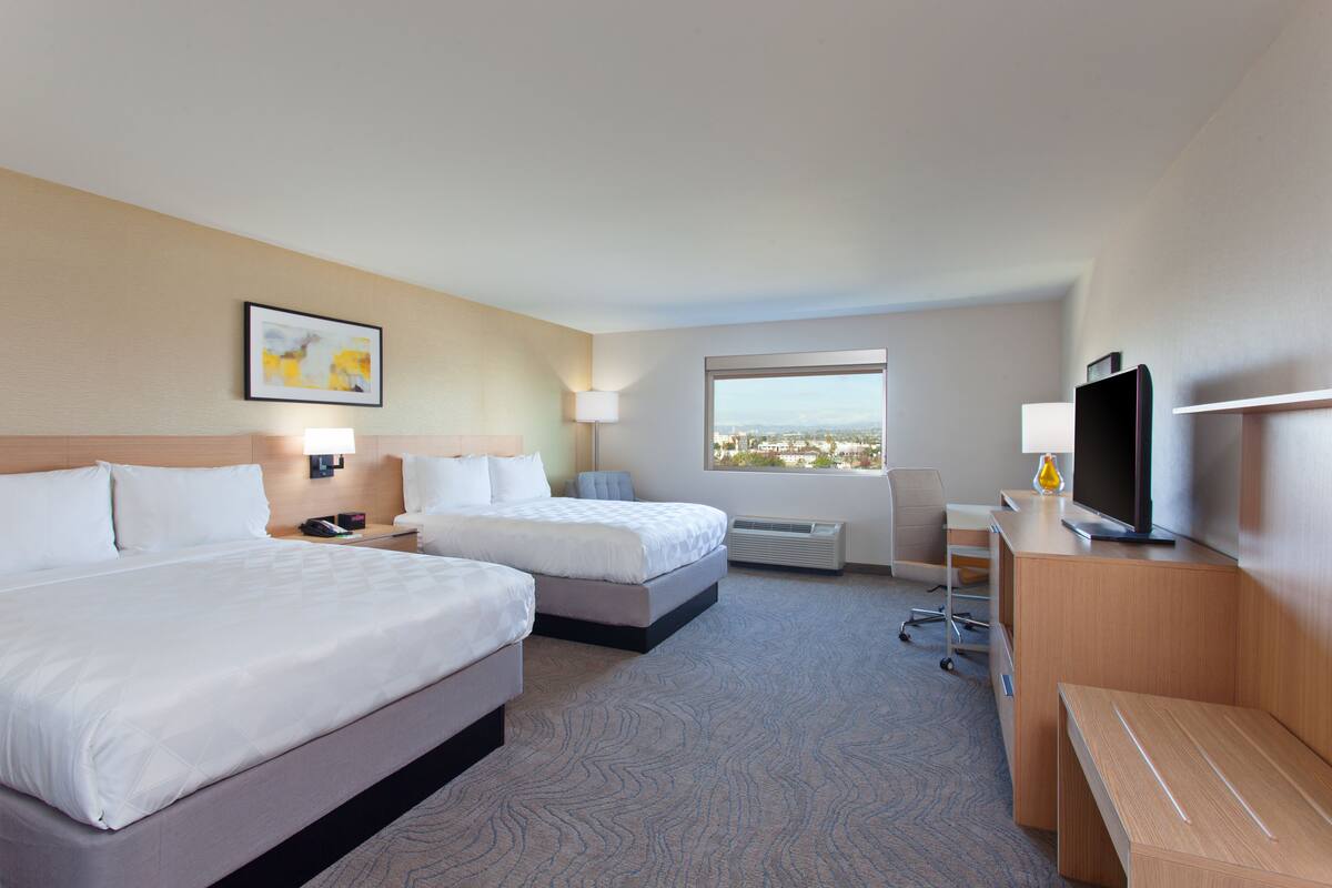 A hotel room with two beds, a desk, a television, and a window with a city view.
