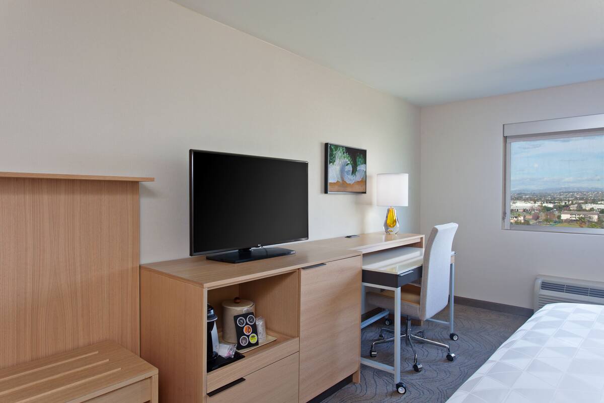A hotel room with a bed, a desk with a computer, a TV, a chair, a window with a city view, and a painting on the wall.