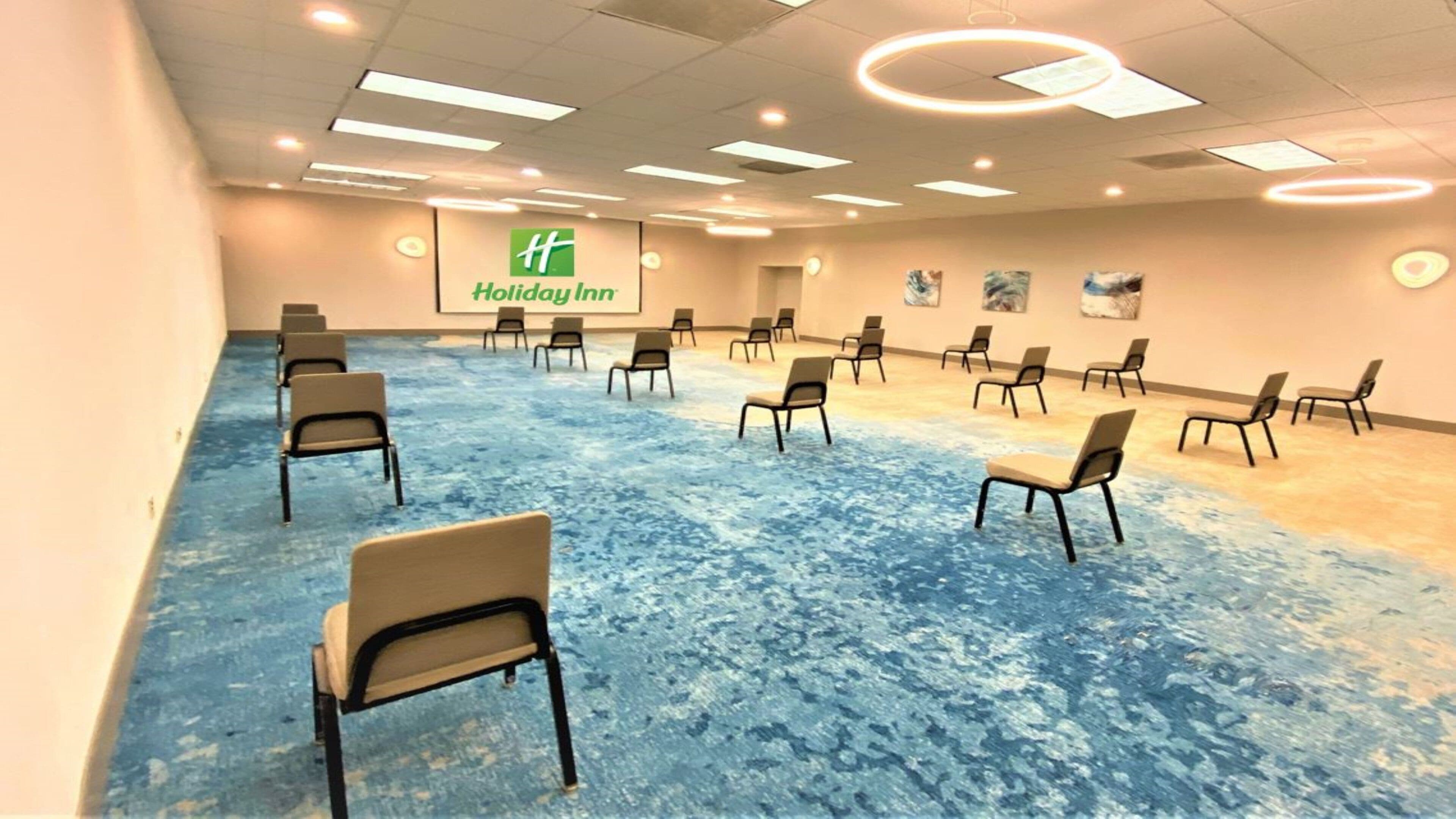 Une salle de conférence dotée d’un tapis bleu, de rangées de chaises, d’un écran de projection arborant le logo « Holiday Inn » et d’un plafond équipé d’un éclairage encastré.