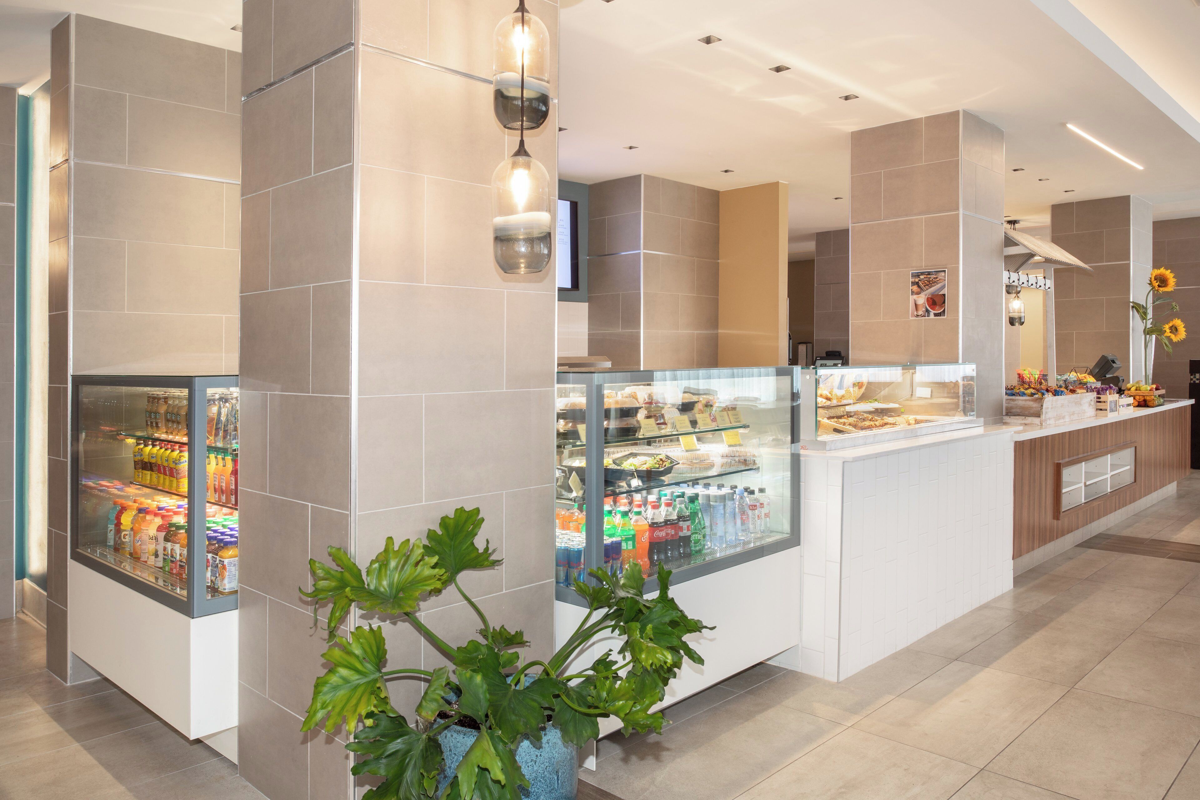 Um hall de hotel moderno com área de café da manhã por autoatendimento, incluindo refrigeradores de bebidas e diversas opções de alimentos.