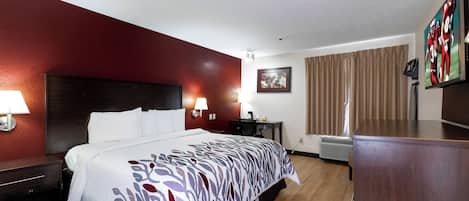 A hotel room with a bed, bedside tables, a desk, a chair, a lamp, a picture frame, a television, and a window with curtains.