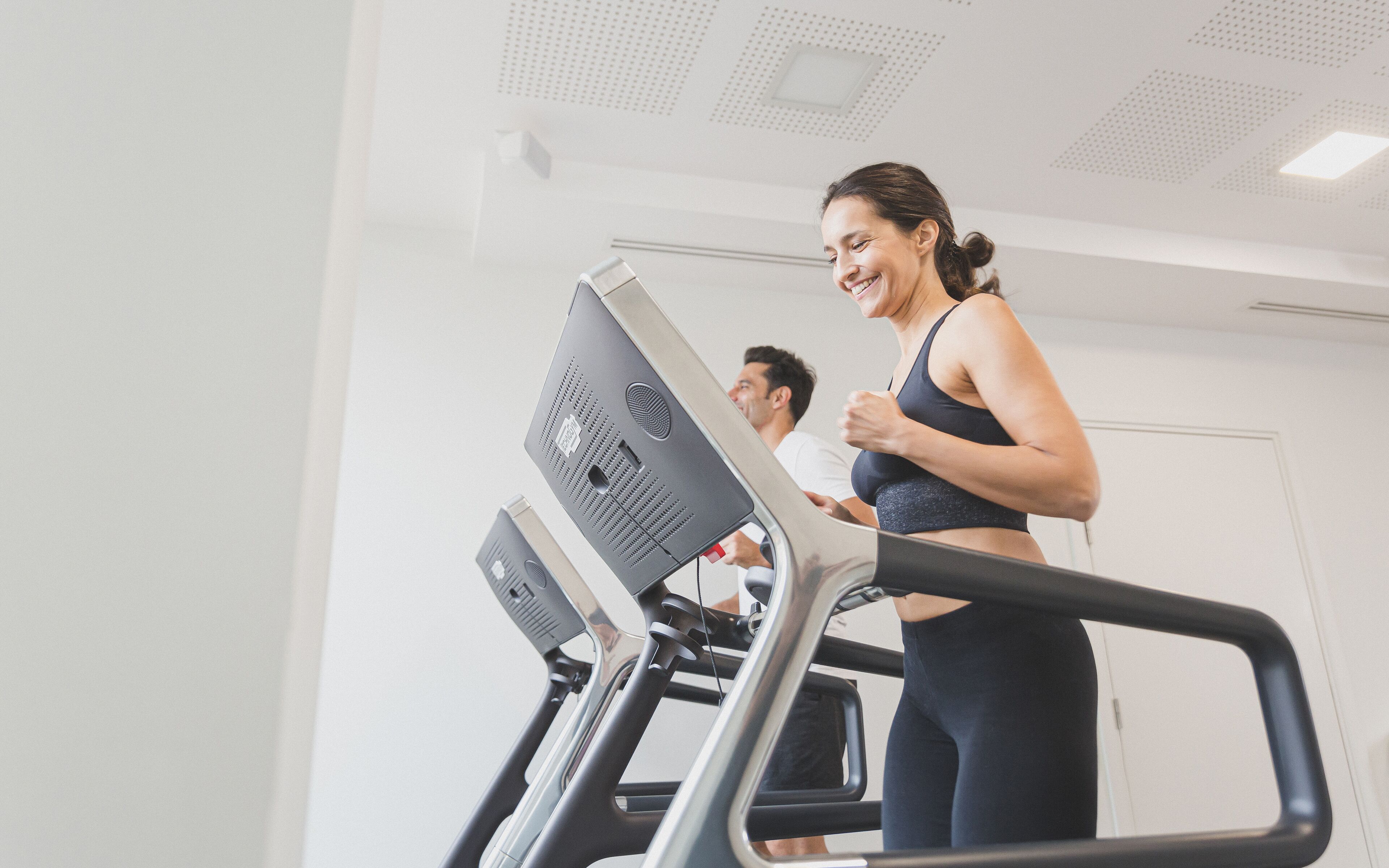 A woman running on a treadmill in a gym with a man in the background.