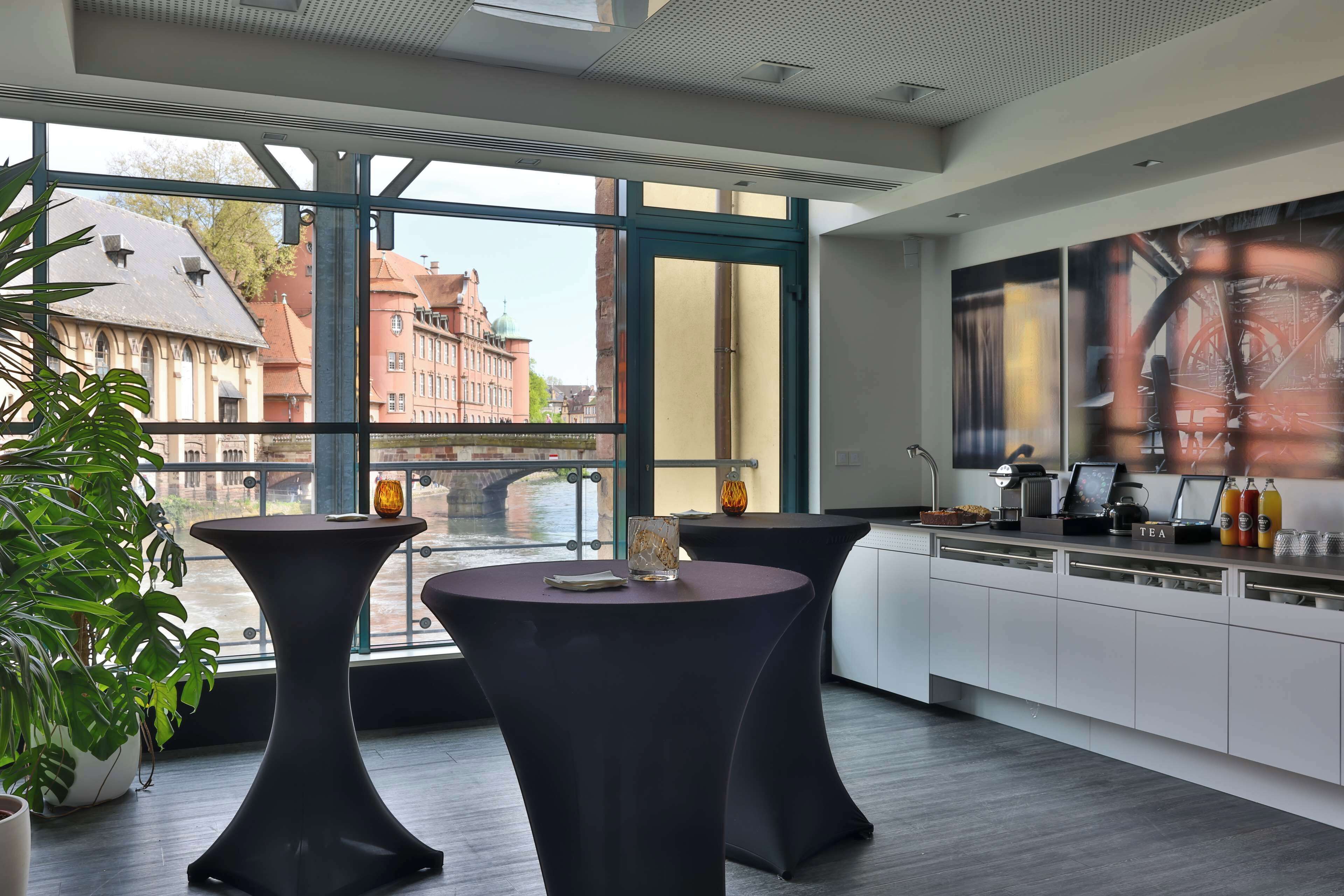 Un intérieur moderne avec deux tables rondes, un coin cuisine et de grandes fenêtres donnant sur des immeubles et une rivière.