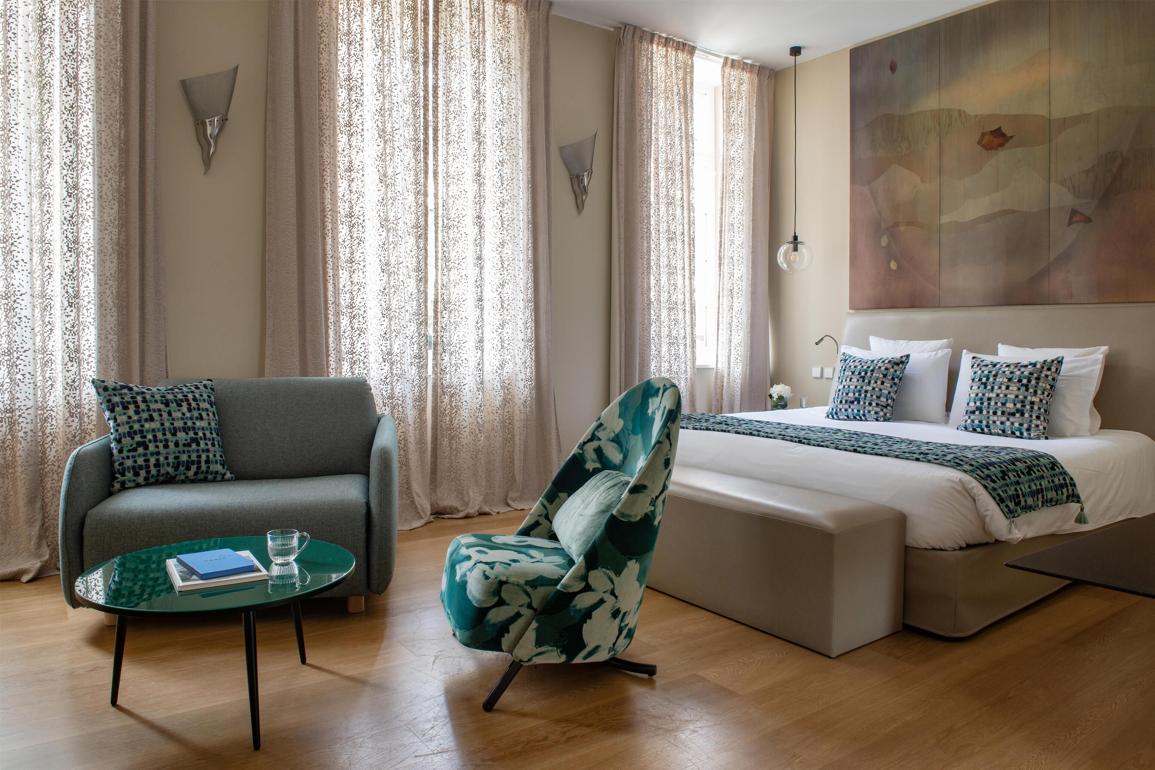A hotel room with a sofa, armchair, and a bed with patterned pillows.
