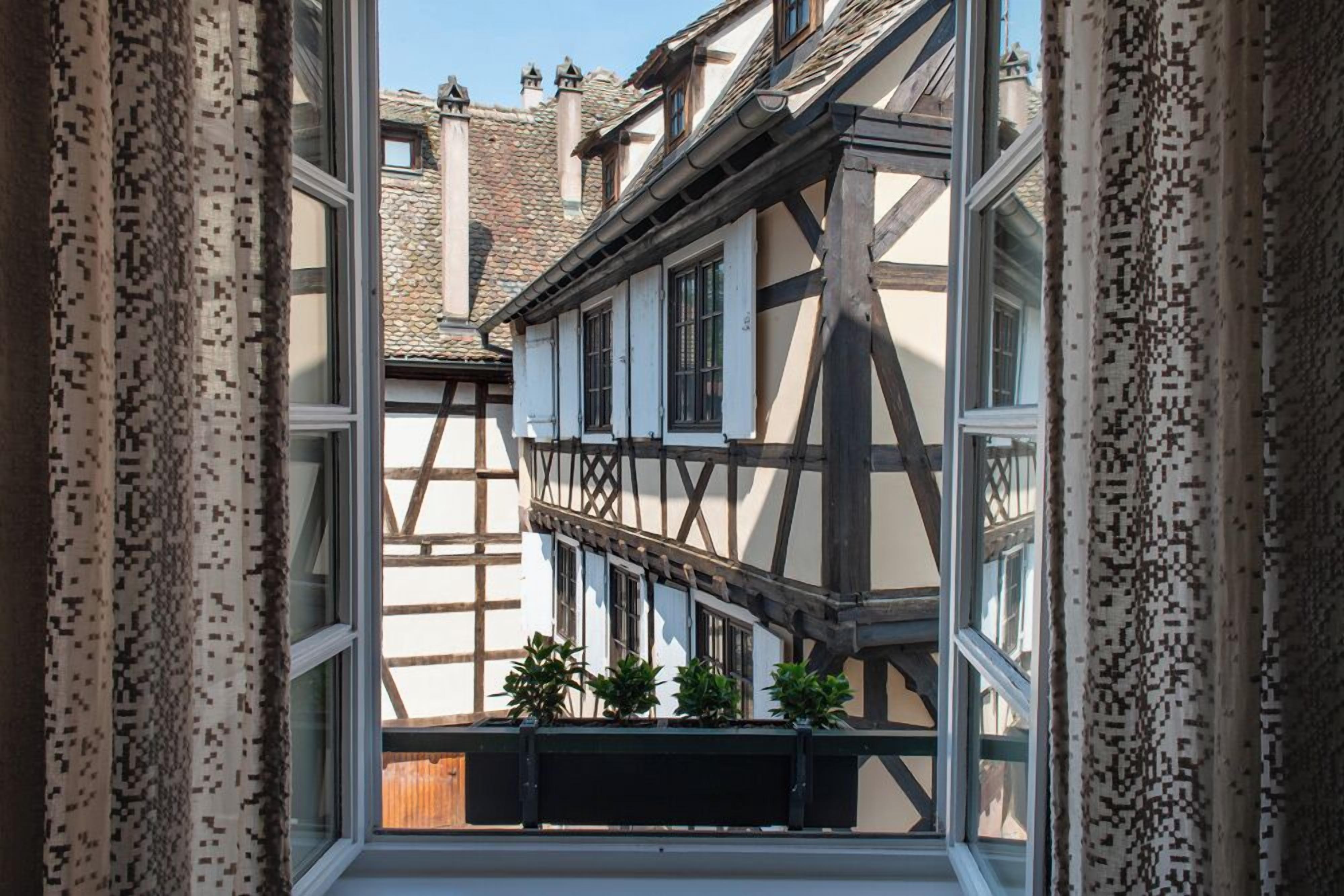 A narrow alleyway with traditional half-timbered buildings, a window with curtains, and a small balcony with potted plants.