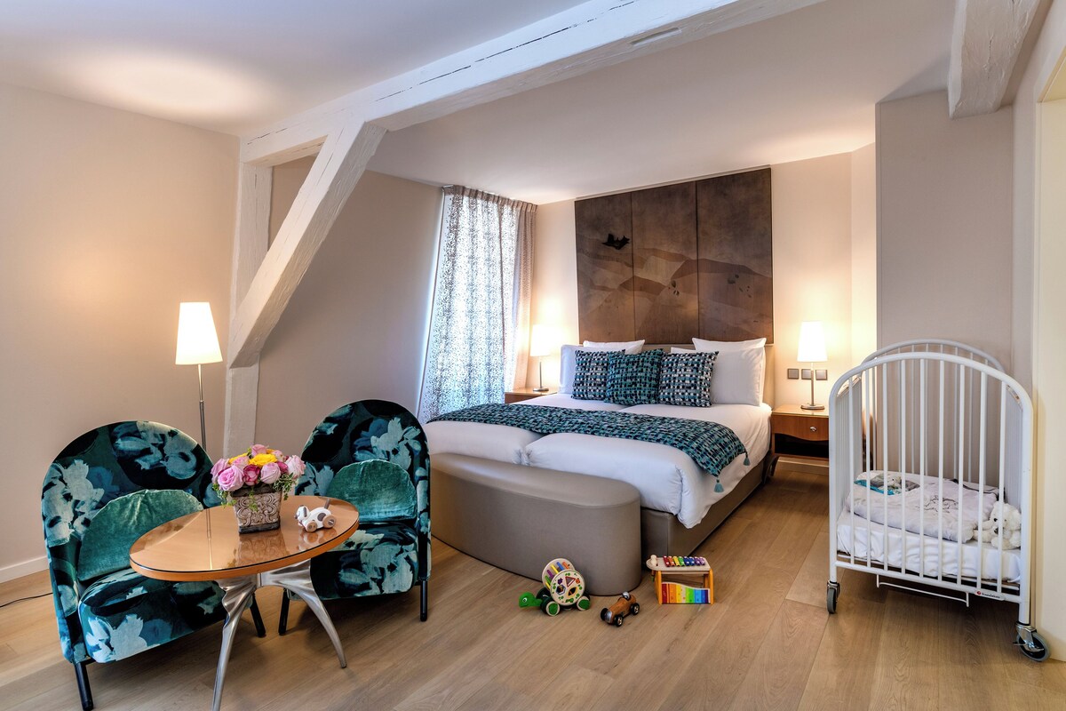 A bedroom with a bed, a crib, a table with a vase, and two chairs.