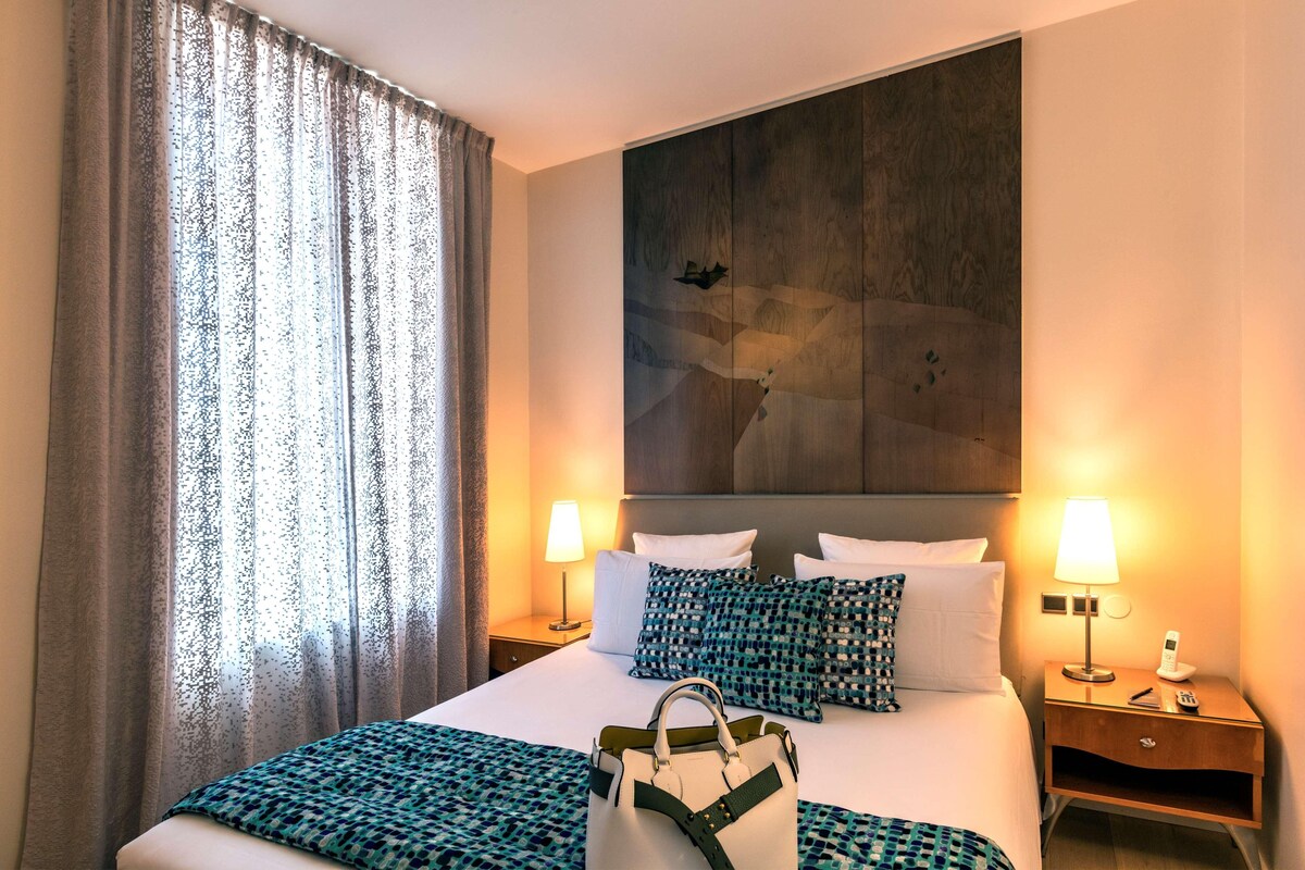A hotel room with a large bed, two bedside tables with lamps, a wall-mounted artwork, and a large window with curtains.