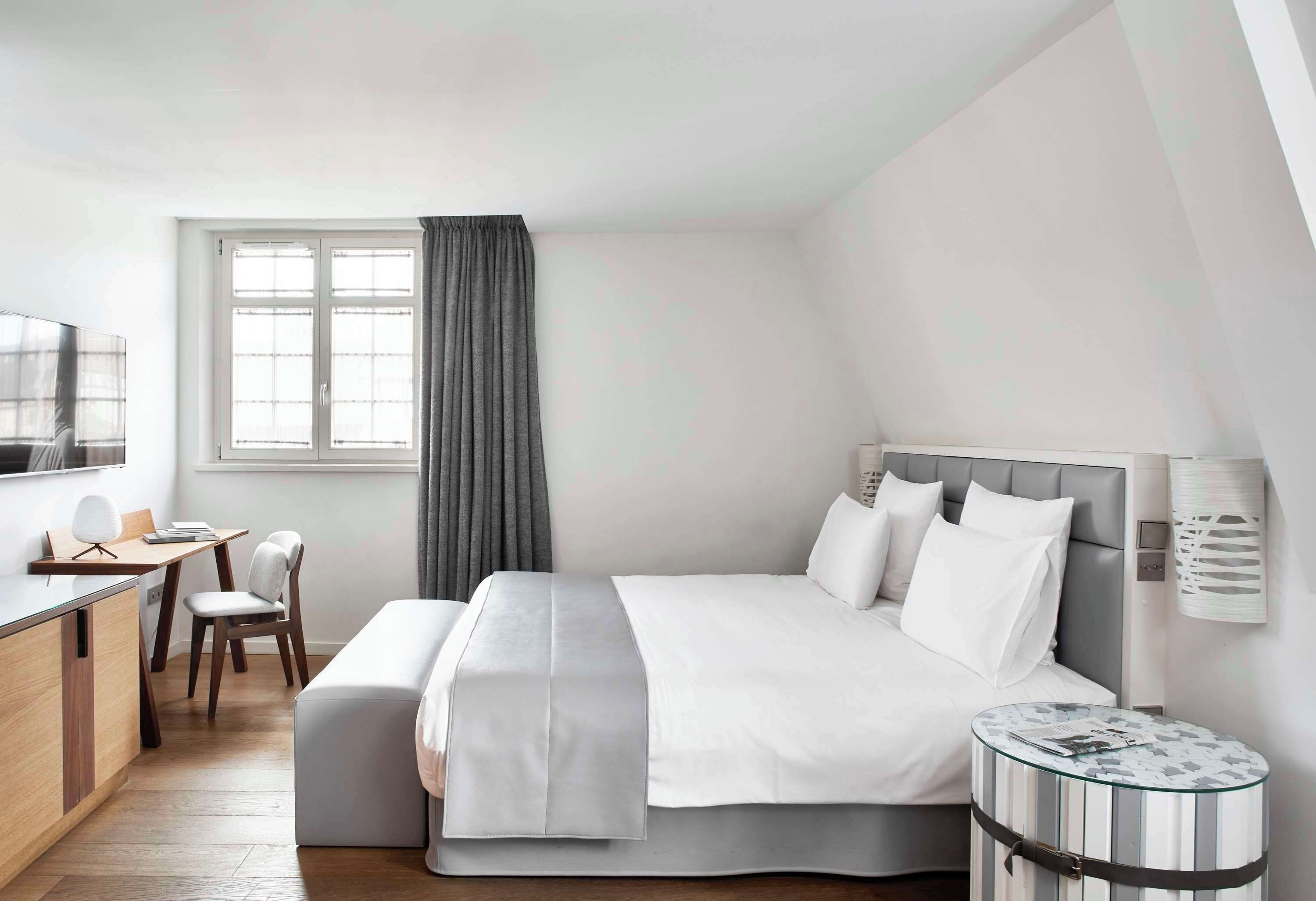 A modern hotel room with a large bed, a desk, a chair, and a small round table.