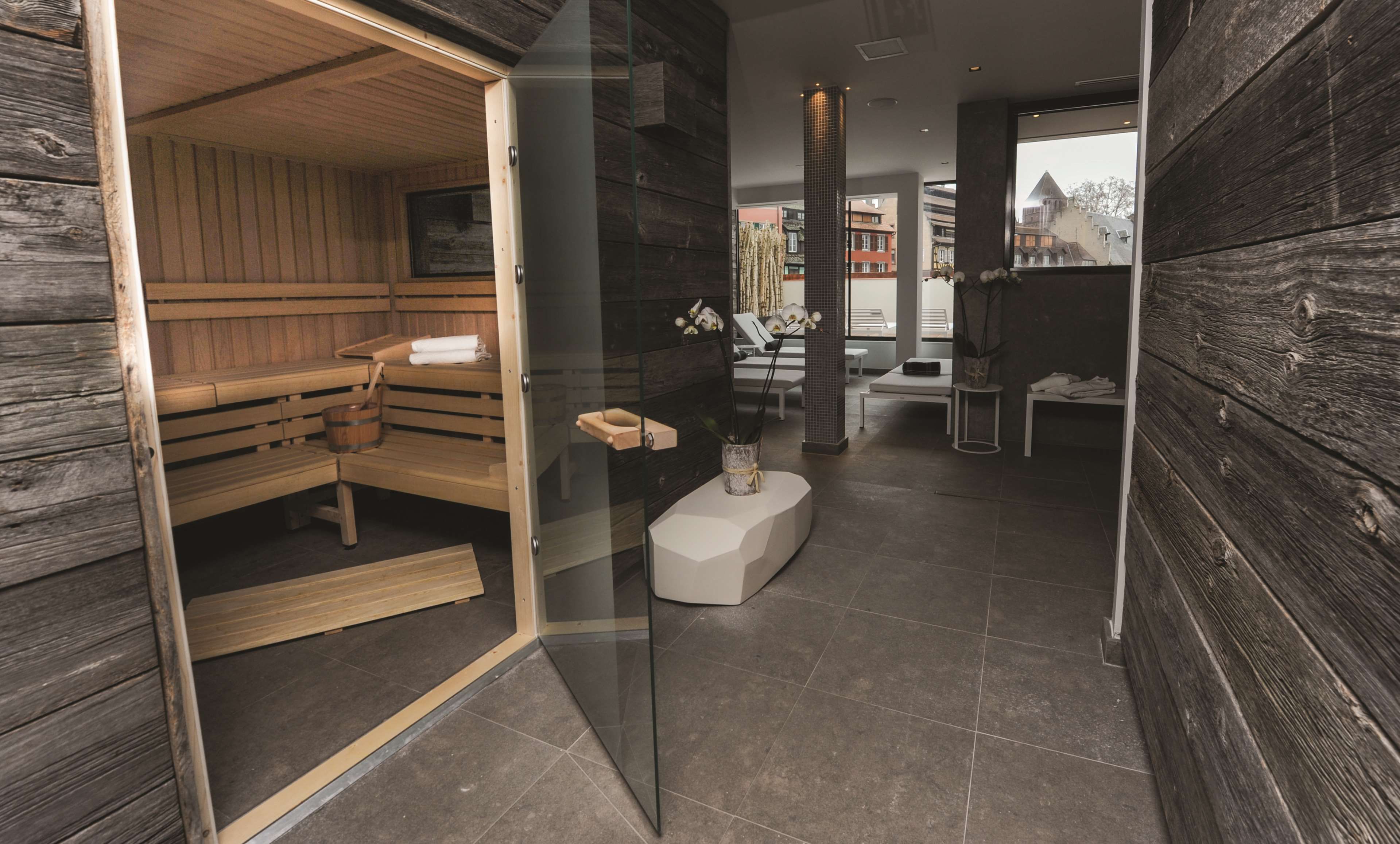 A sauna room with wooden benches and a glass door leading to a relaxation area with a bench and a small table.