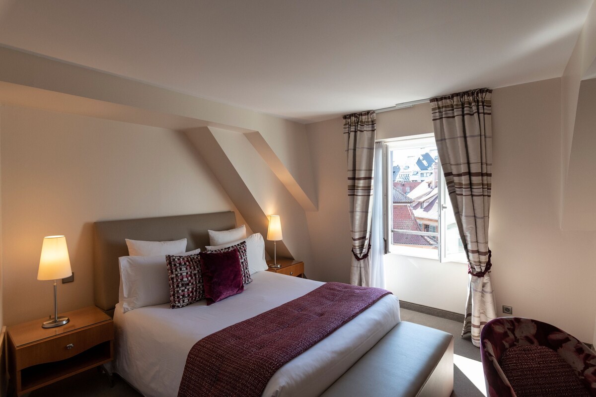 Un dormitorio con cama, mesitas de noche, una silla, ventana con cortinas y vistas a edificios.