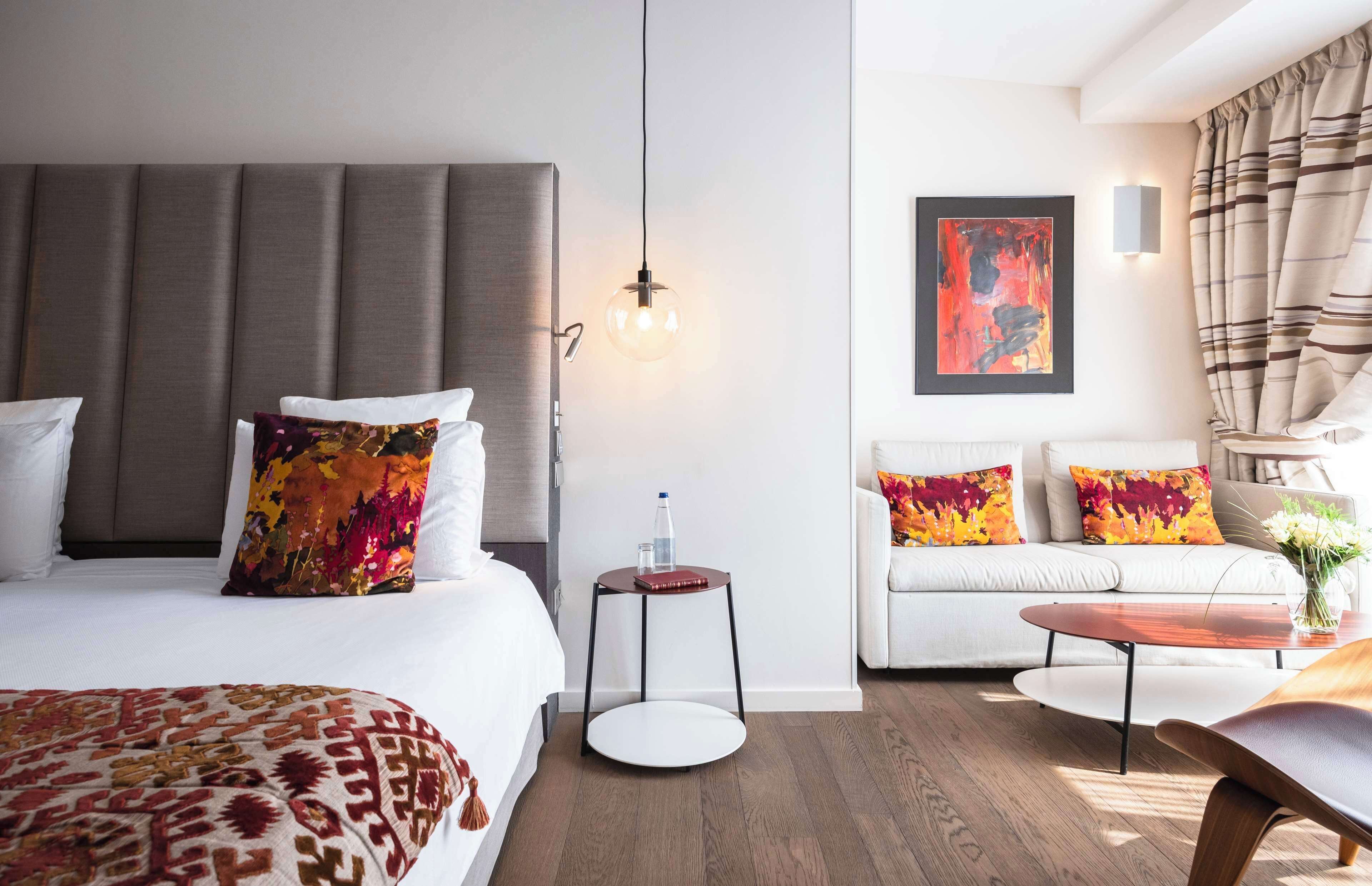 A modern hotel room with a large bed, a sofa, a small table, and a hanging light fixture.
