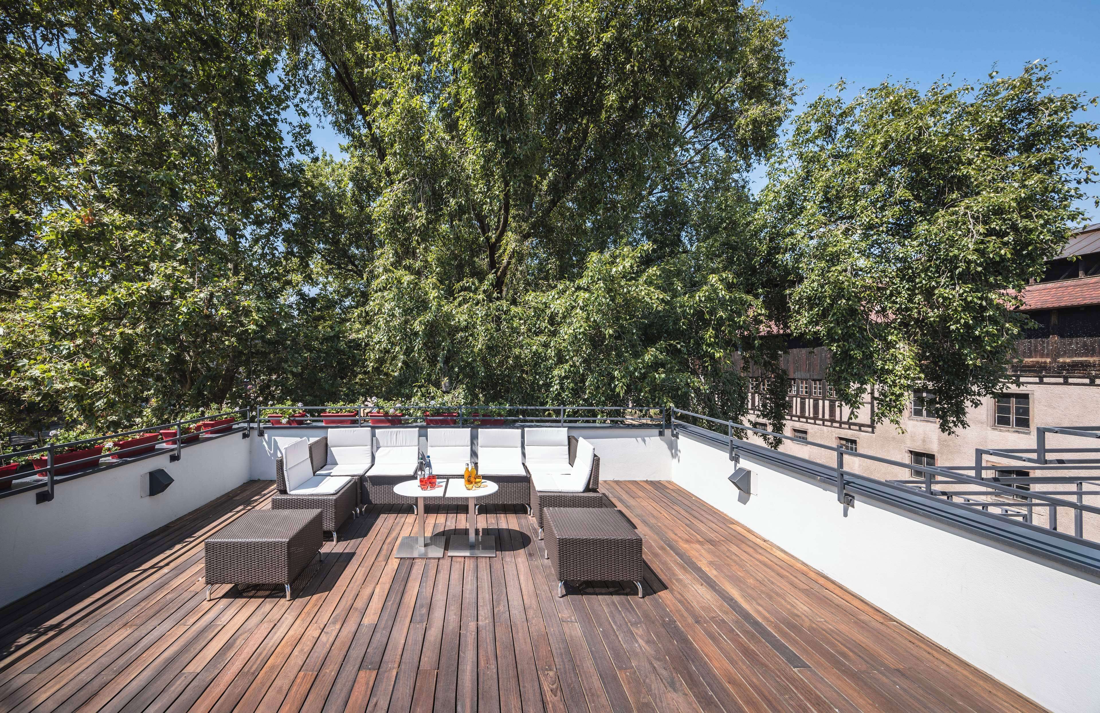 Una terraza en la azotea con piso de madera, mobiliario exterior y vistas a árboles y edificios.