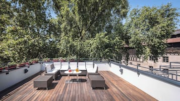Una terraza en la azotea con piso de madera, mobiliario exterior y vistas a árboles y edificios.