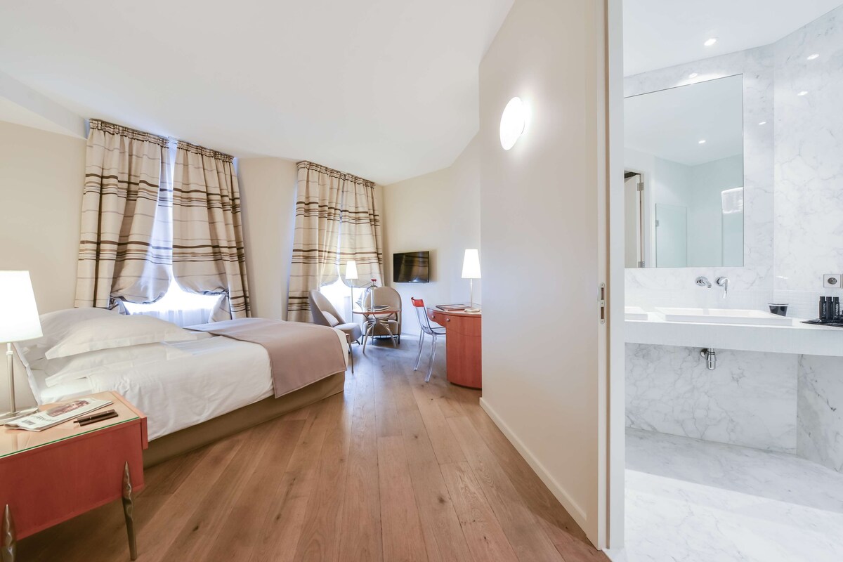 Habitación de hotel con cama, mesita de noche, silla, escritorio y baño con espejo y lavabo.