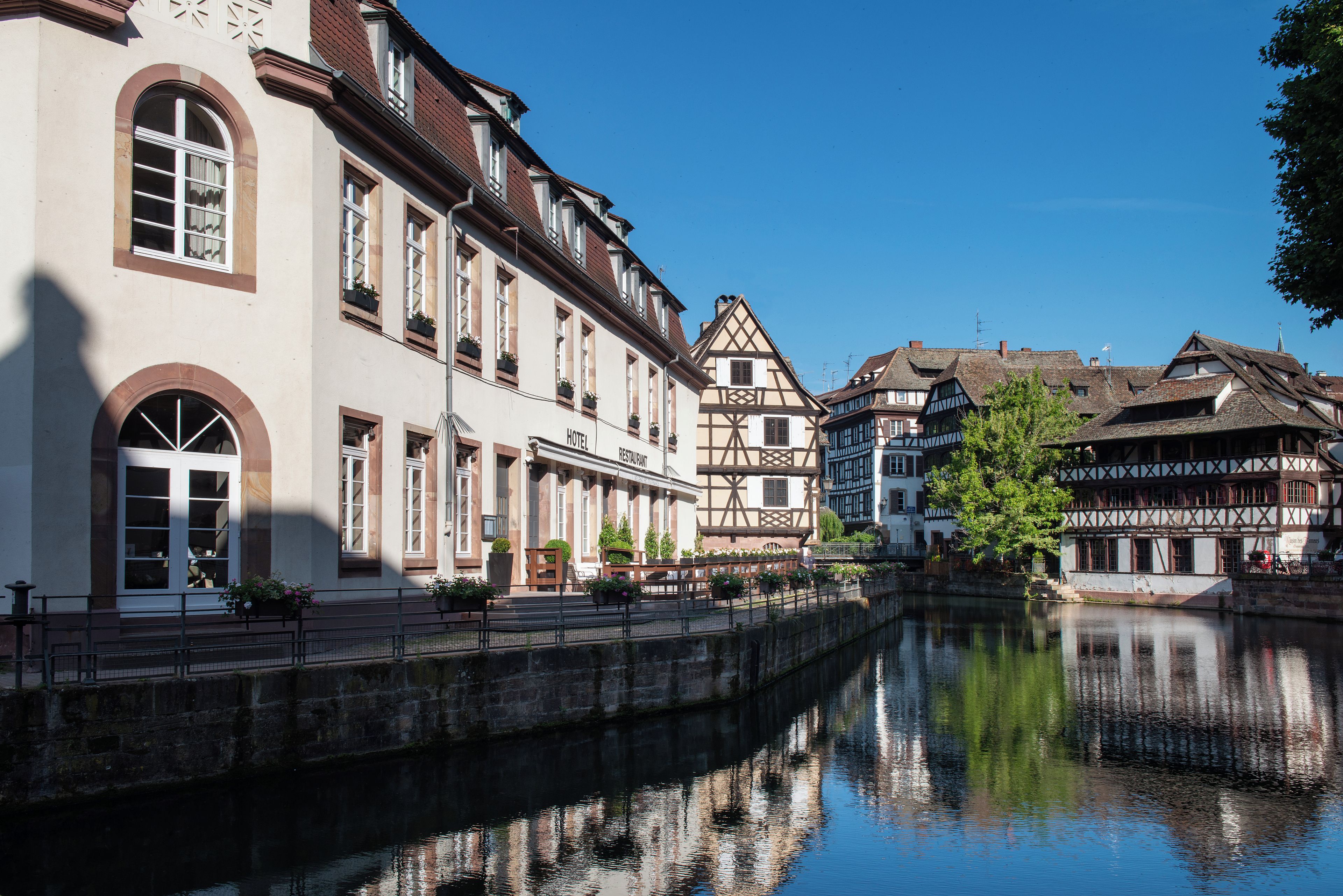 A riverside hotel with a traditional European architectural style, featuring a large arched window and a balcony with potted plants.