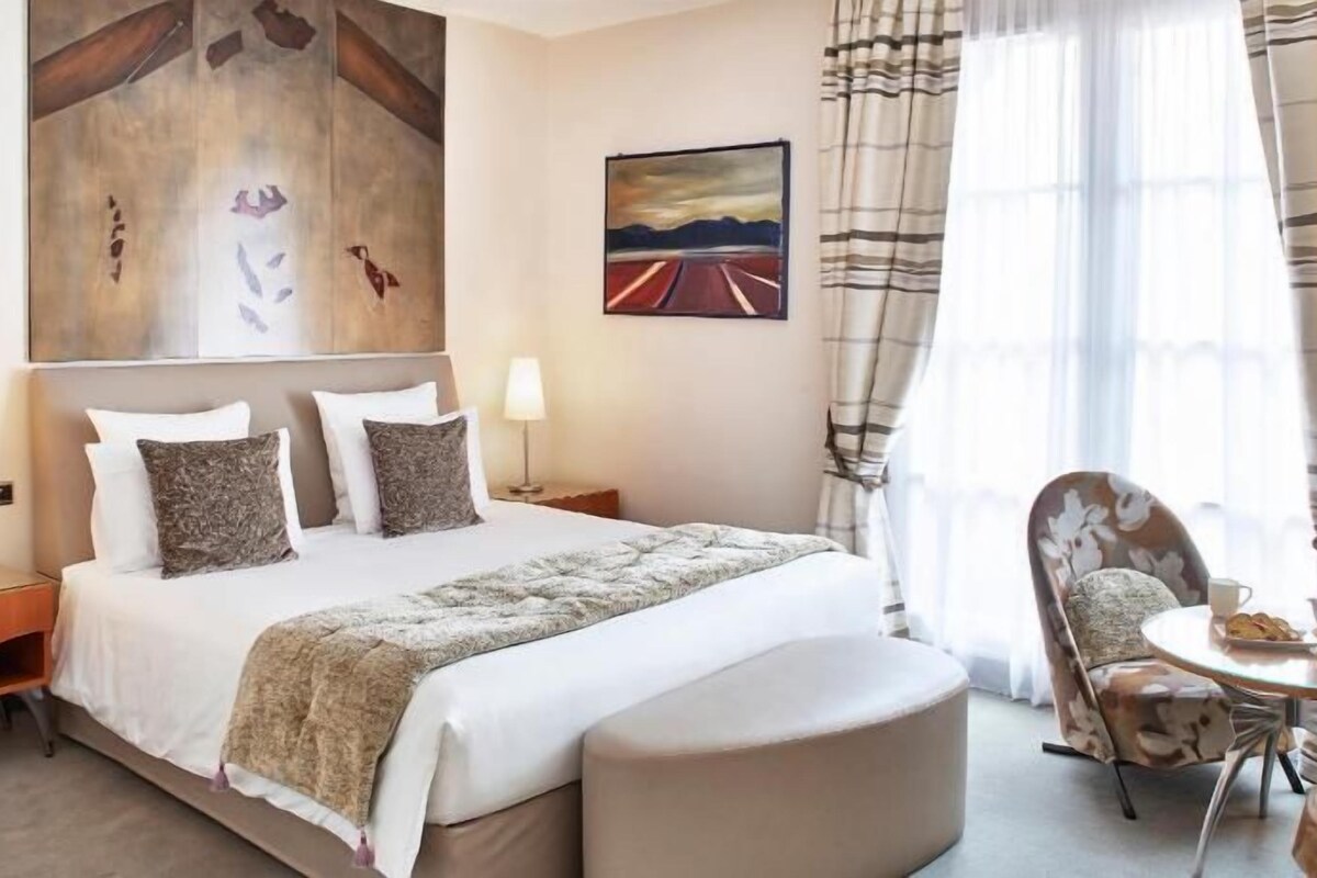 Una habitación de hotel con una cama, dos mesitas de noche, una silla, una pequeña mesa con una taza y pasteles, y dos cuadros en las paredes.