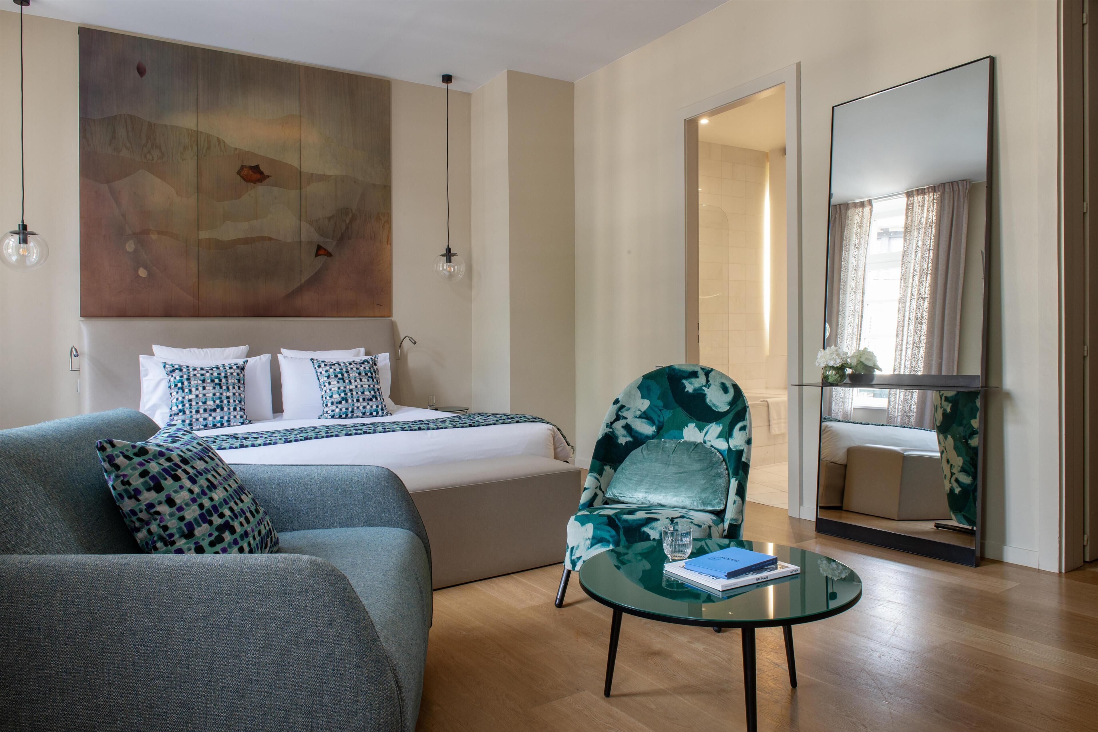 Een moderne hotelkamer met een bed, een slaapbank, een ronde glazen tafel en een grote spiegel.