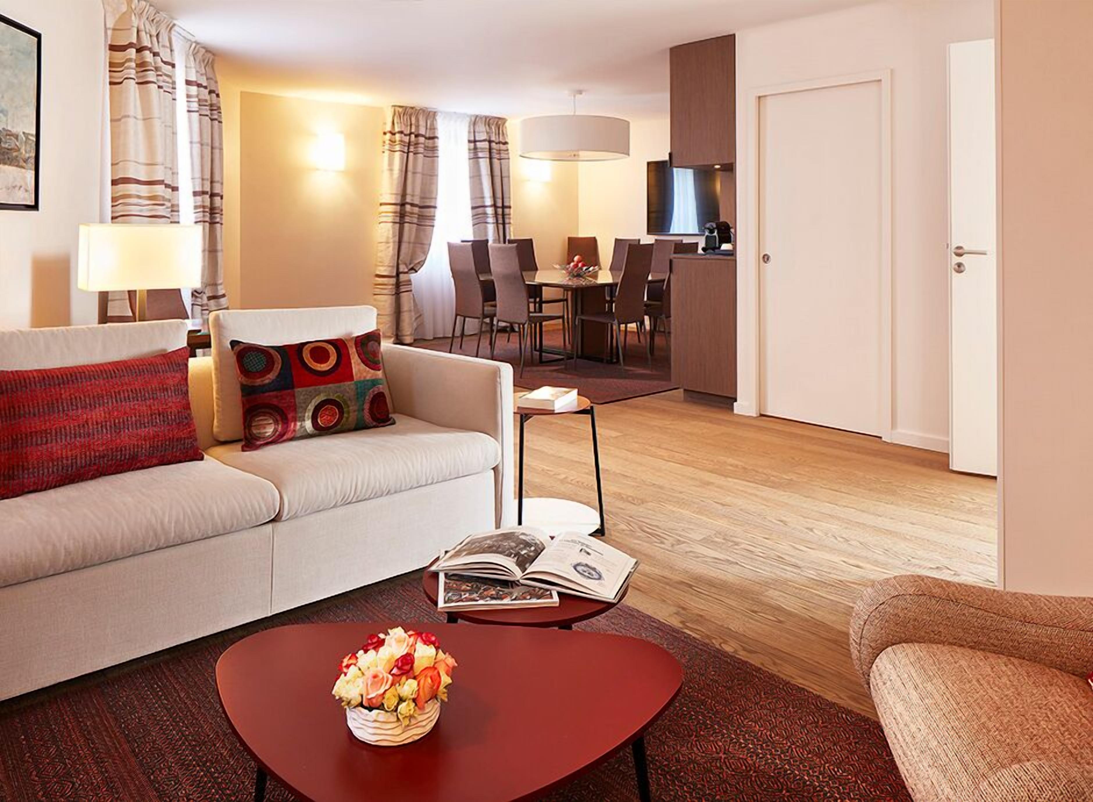 Un salon moderne comprenant un canapé, une table basse avec un panier de fruits, un coin repas avec une table et des chaises, et un coin cuisine avec un téléviseur.
