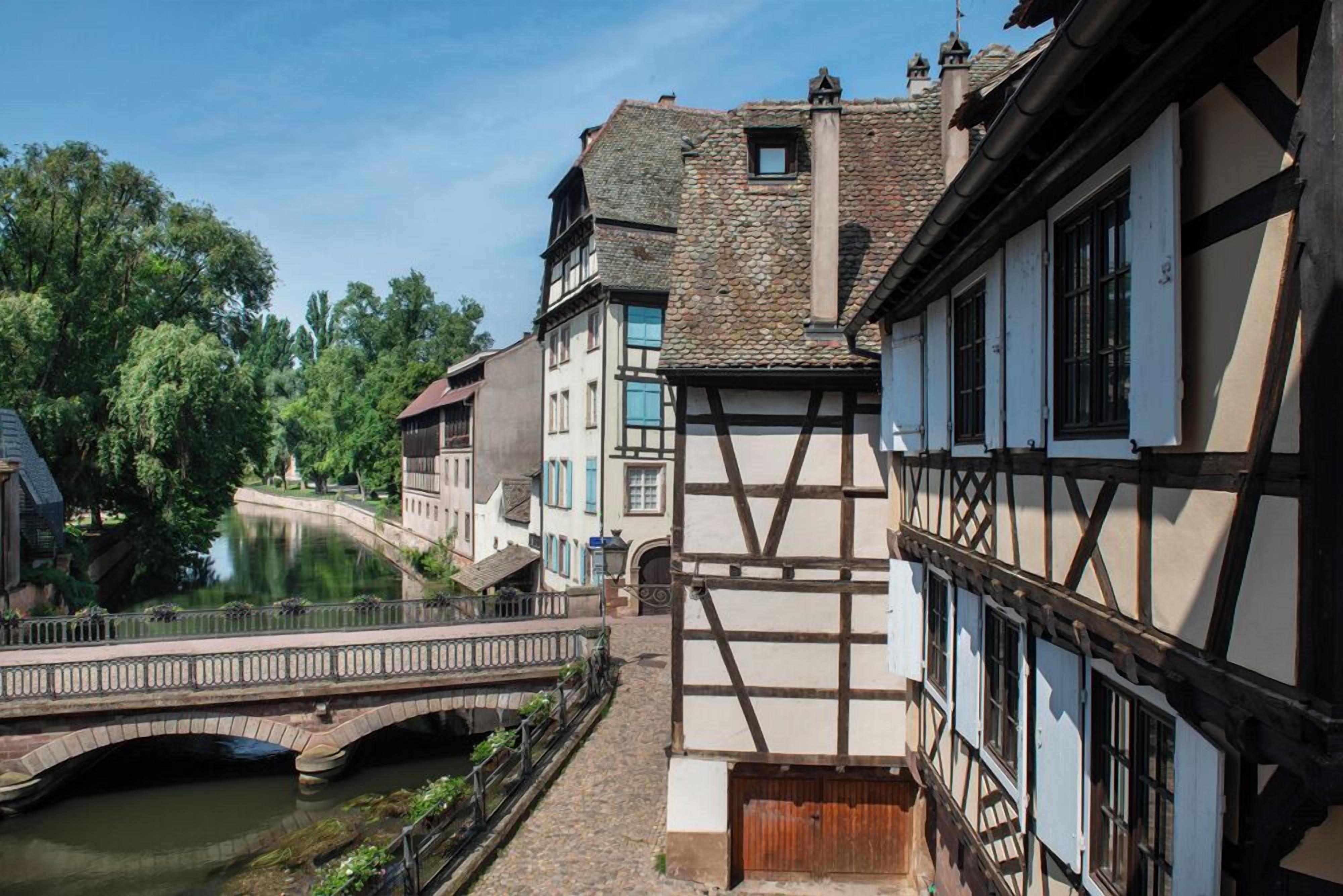 A traditional European town with a stone bridge over a canal, half-timbered buildings, and a clear sky.