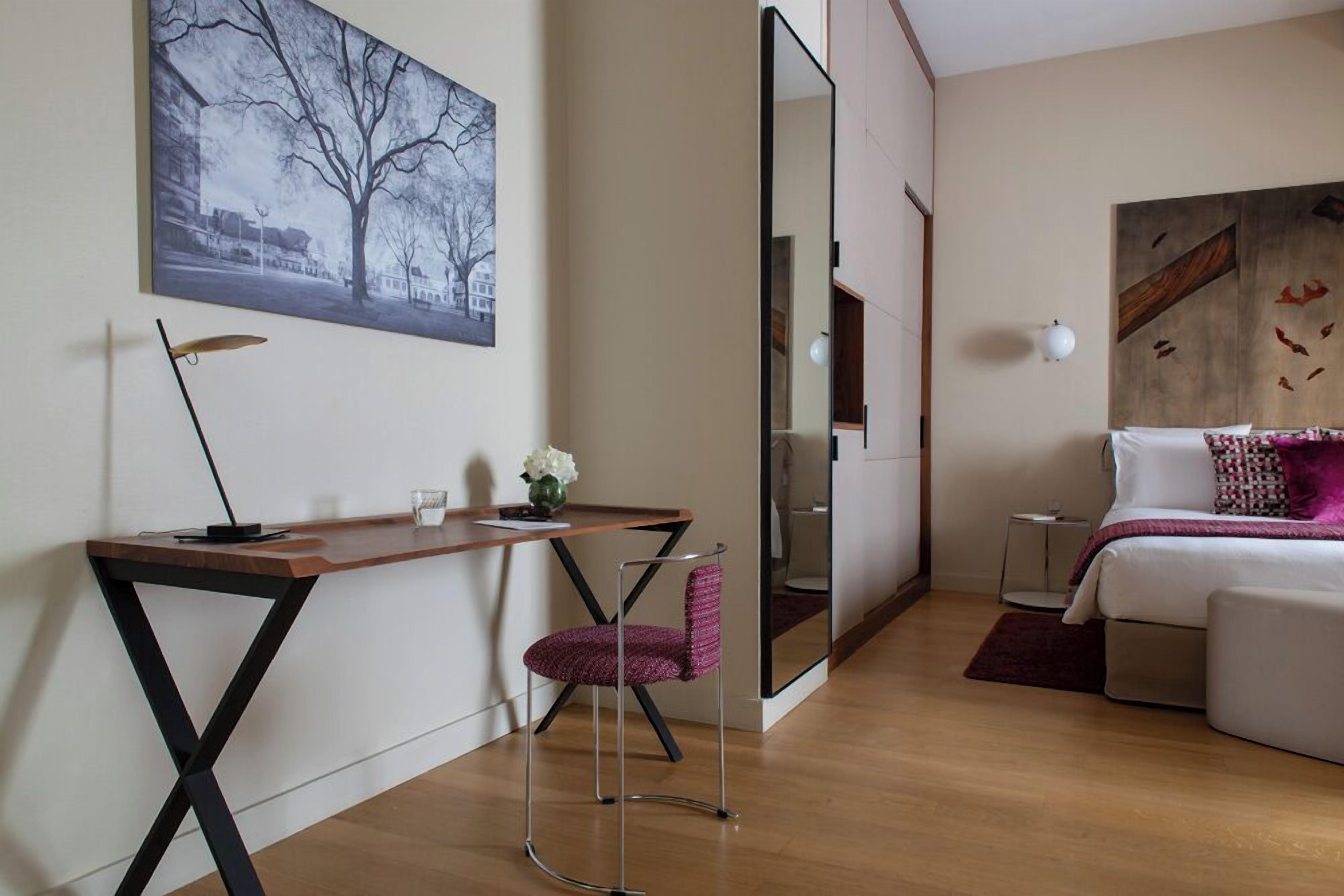 A modern hotel room with a wooden desk, a pink chair, a bed with a white and purple pillow, and a large mirror.