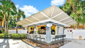 A small outdoor bar with a covered seating area, a counter with various items, and a white fence in the background. - Mi Casa Hotel (Orlando)