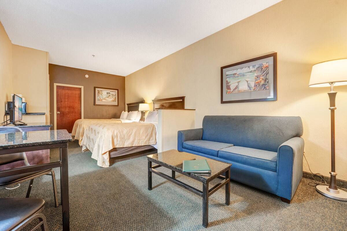 A hotel room with a bed, a blue sofa, a desk with a computer, a coffee table, and a lamp.