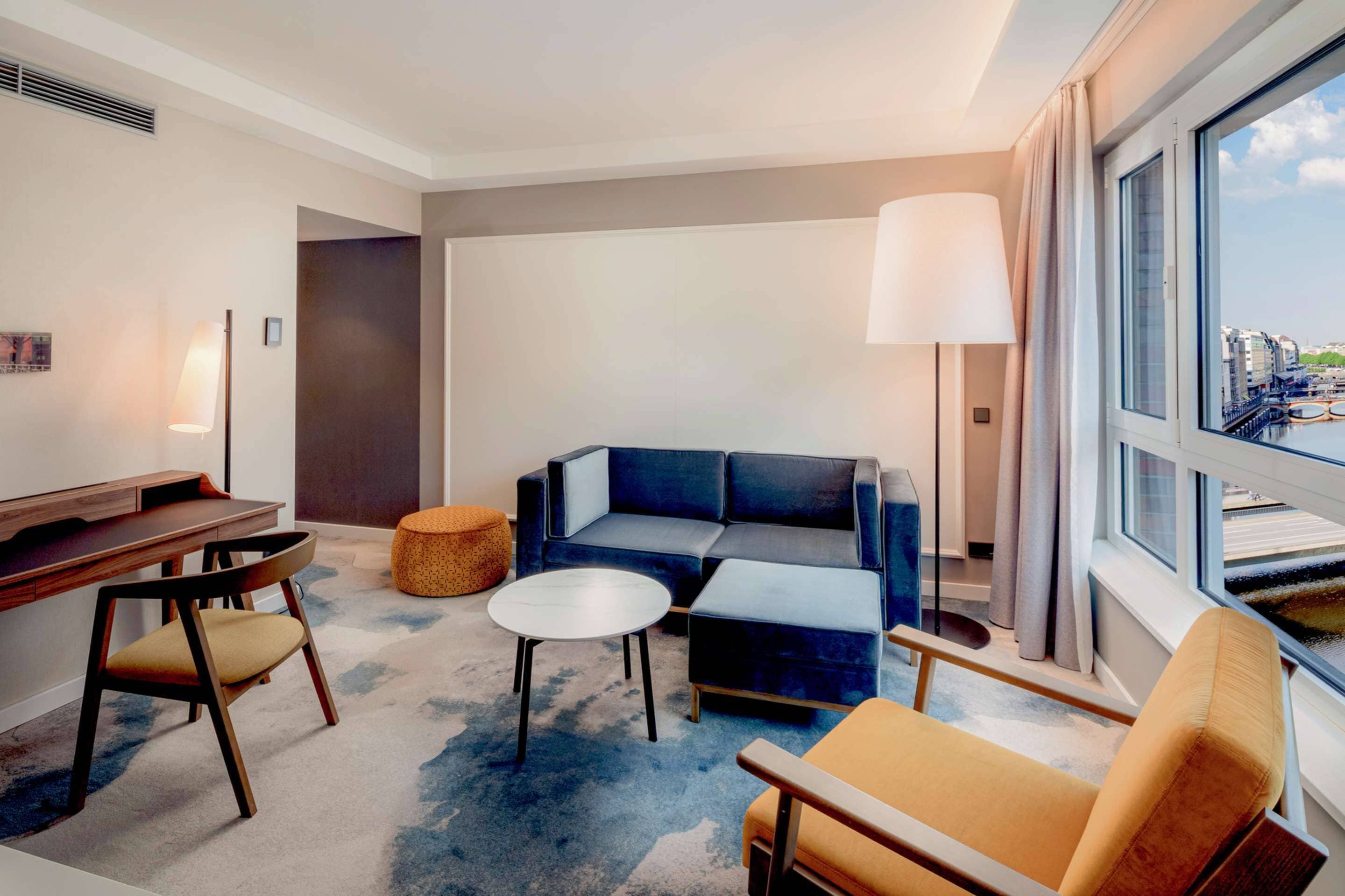 Une chambre d’hôtel moderne équipée d’un canapé, d’un fauteuil, d’un pouf et d’un bureau avec une chaise.
