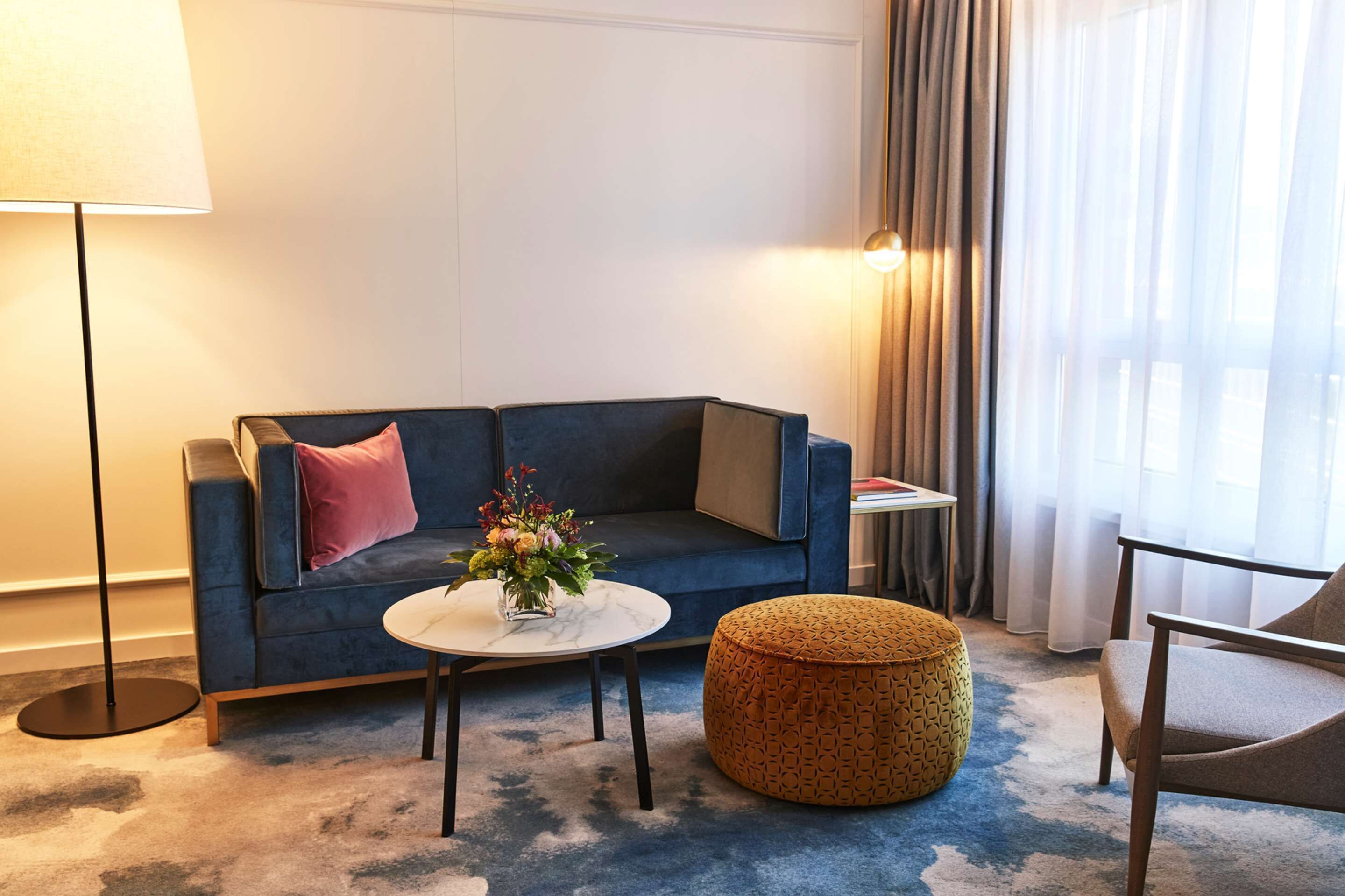 Una sala de estar moderna con un sofá azul, un otoman redondo, una mesita redonda con un jarrón de flores, una lámpara de pie y una silla.