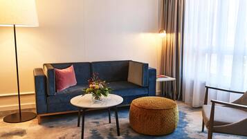 A modern living room with a blue sofa, a round ottoman, a small round table with a vase of flowers, a floor lamp, and a chair.
