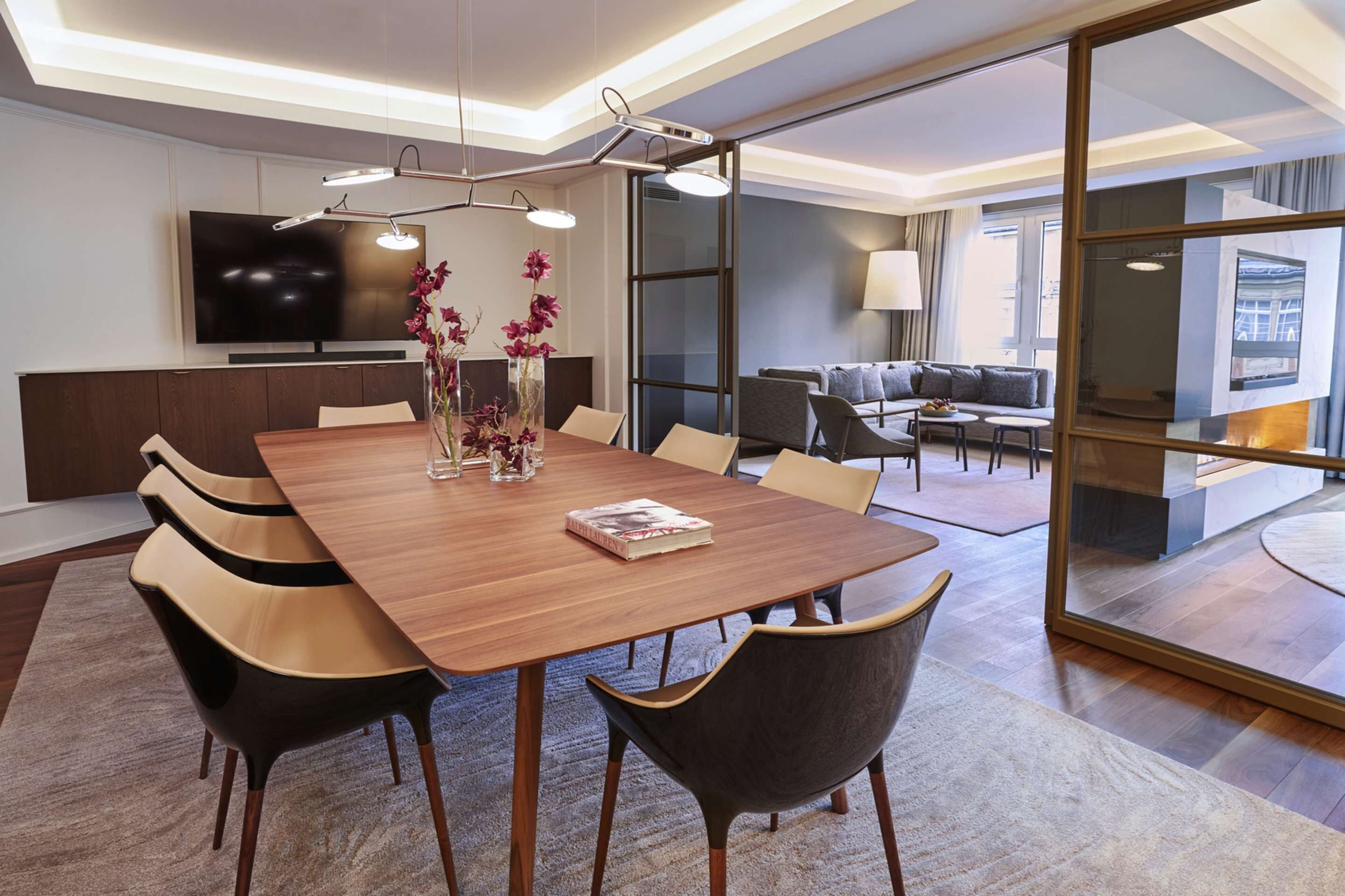 A modern hotel room with a wooden dining table, comfortable seating, a flat-screen TV, and a large window offering a view of the city.
