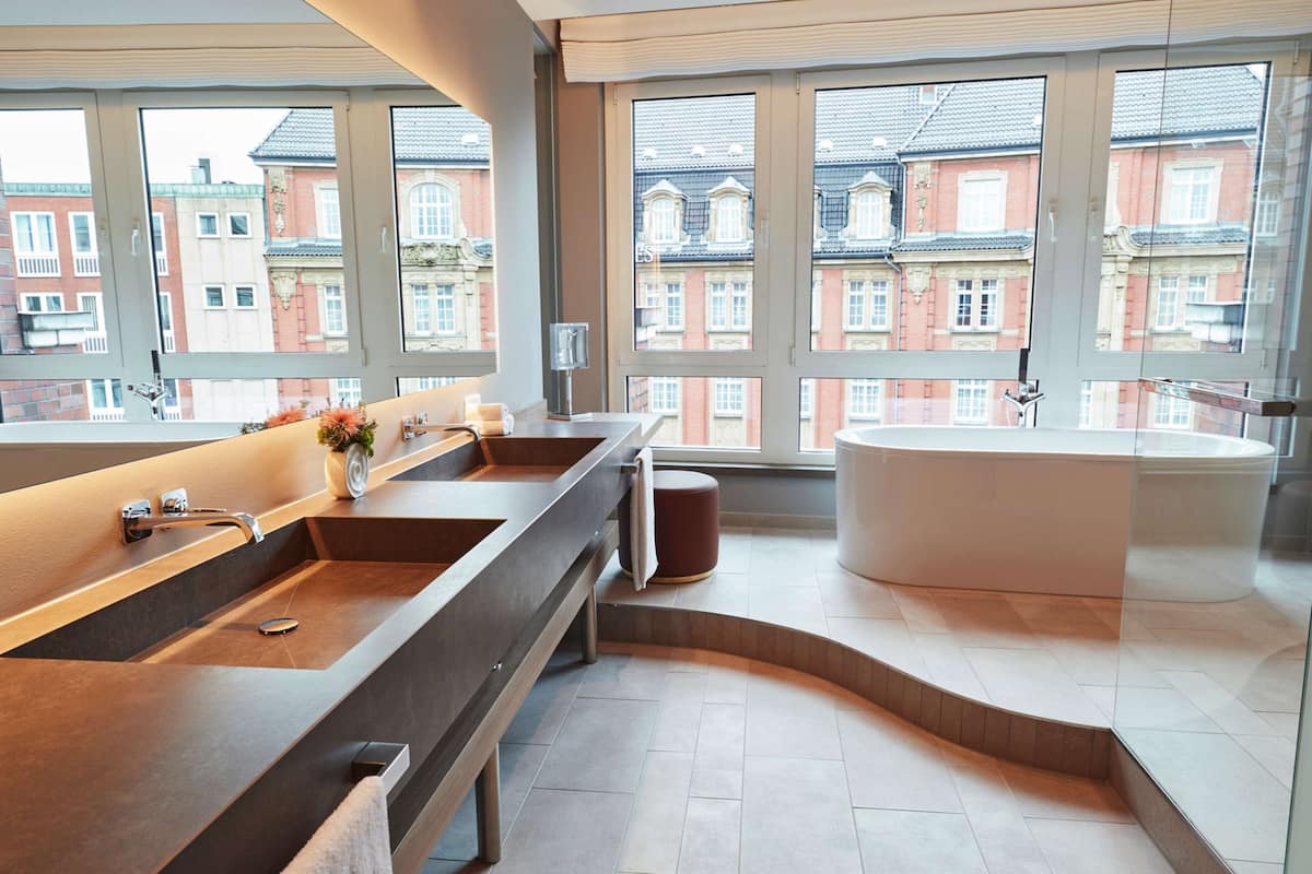 Une salle de bain moderne dotée d’un grand lavabo, d’une baignoire et d’une fenêtre donnant sur des immeubles aux façades en briques rouges.