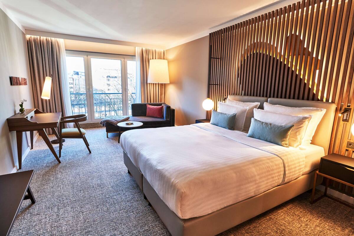 Une chambre d’hôtel avec un grand lit, un bureau avec une chaise, une petite table et une vue sur la ville grâce à de grandes fenêtres.