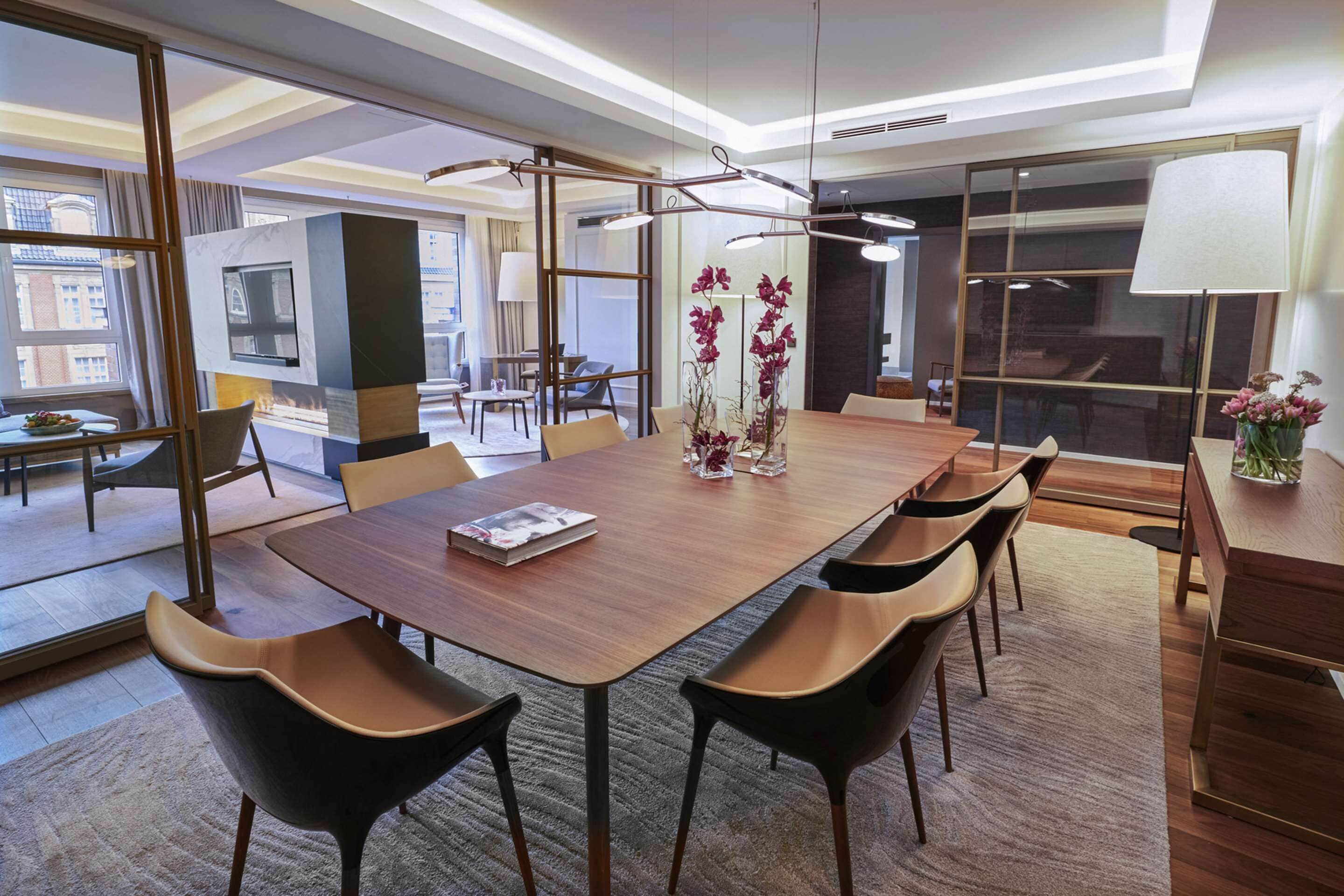 A modern hotel dining area with a long wooden table, comfortable chairs, and a central vase with flowers.