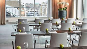 A hotel conference room with tables set for meetings, large windows with a city view, and a large potted plant on one side.