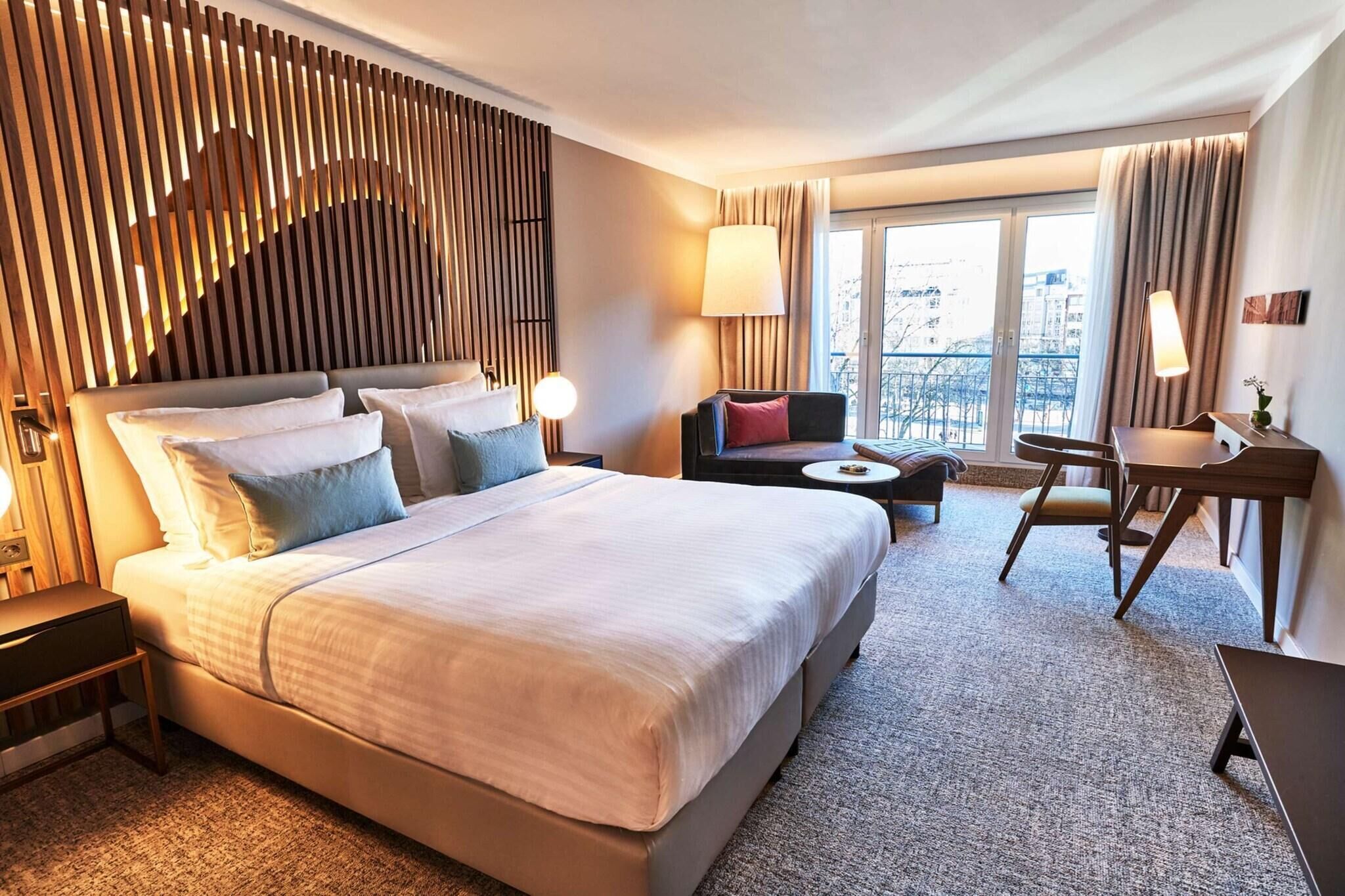 Ein Hotelzimmer mit einem großen Bett, einem Schreibtisch, einem Sessel und einer Aussicht auf die Stadt durch das Fenster.