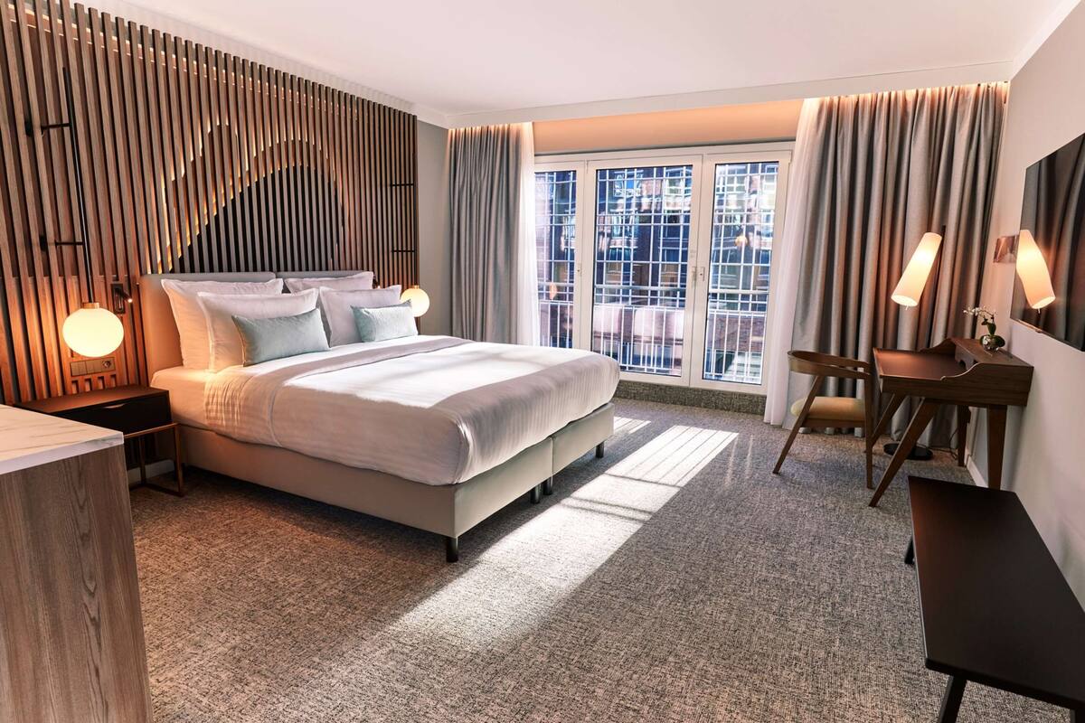 Une chambre d’hôtel moderne, dotée d’un grand lit, d’un bureau et d’une chaise. Une fenêtre, ornée de rideaux, offre une vue sur les bâtiments environnants.