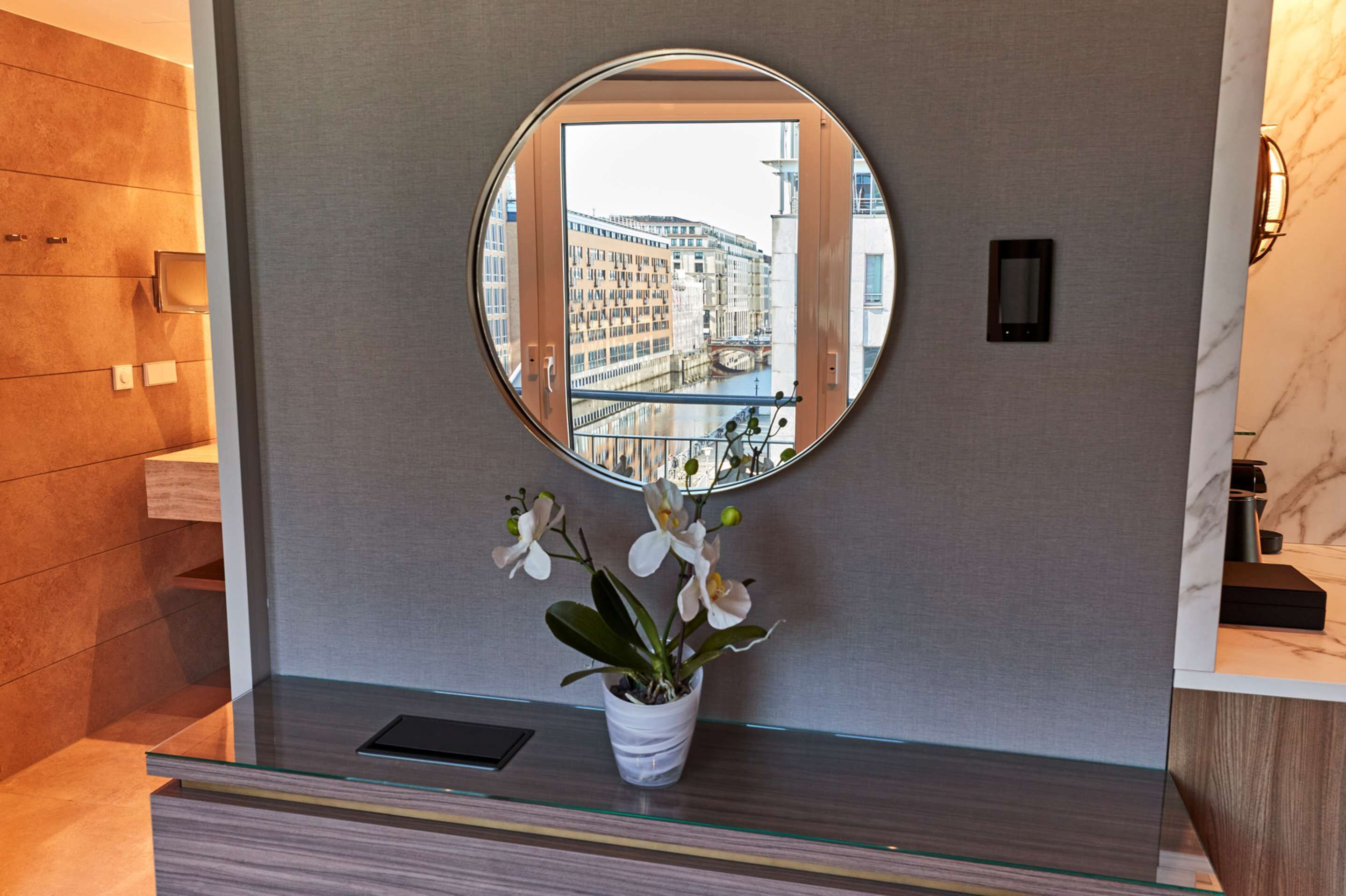 Ein modernes Badezimmer mit einem runden Spiegel, einer Orchidee im Topf und Blick auf ein Gebäude im Spiegel.