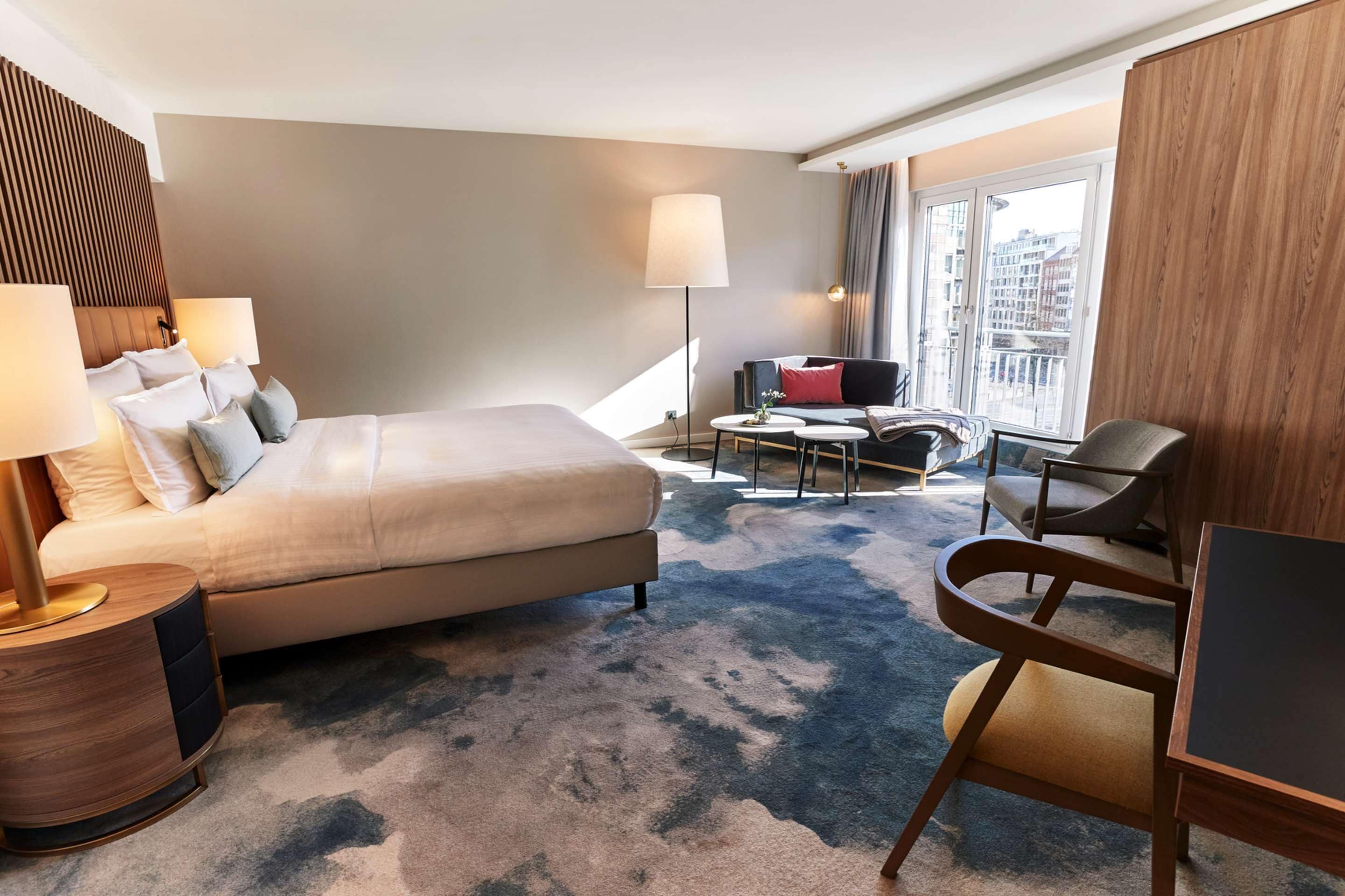 Ein Hotelzimmer mit einem großen Bett, einem Schreibtisch, einem Stuhl, einer Lampe und Blick auf die Stadt durch das Fenster.