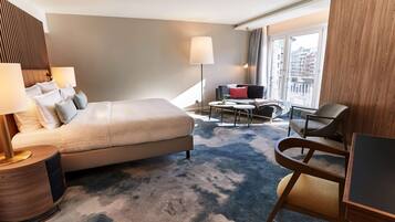 Ein Hotelzimmer mit einem großen Bett, einem Schreibtisch, einem Stuhl, einer Lampe und Blick auf die Stadt durch das Fenster.