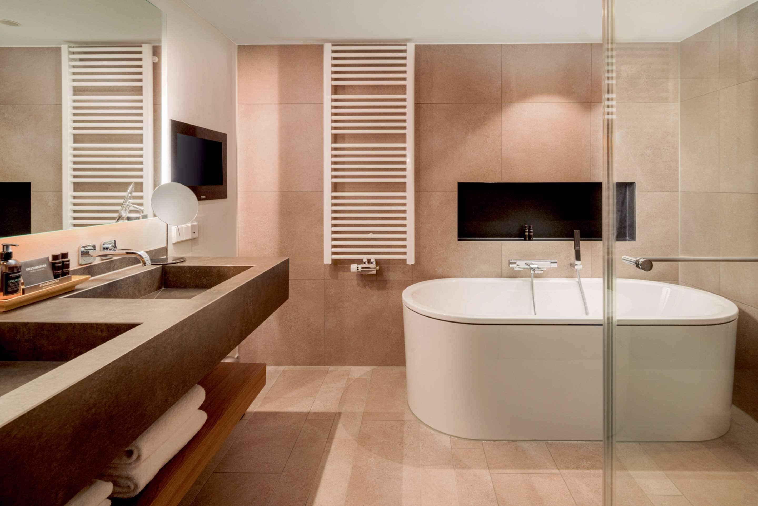 Ein modernes Badezimmer mit einer freistehenden Badewanne, einem großzügigen Waschtisch mit Spiegel und zwei an der Wand montierten Fernsehern.