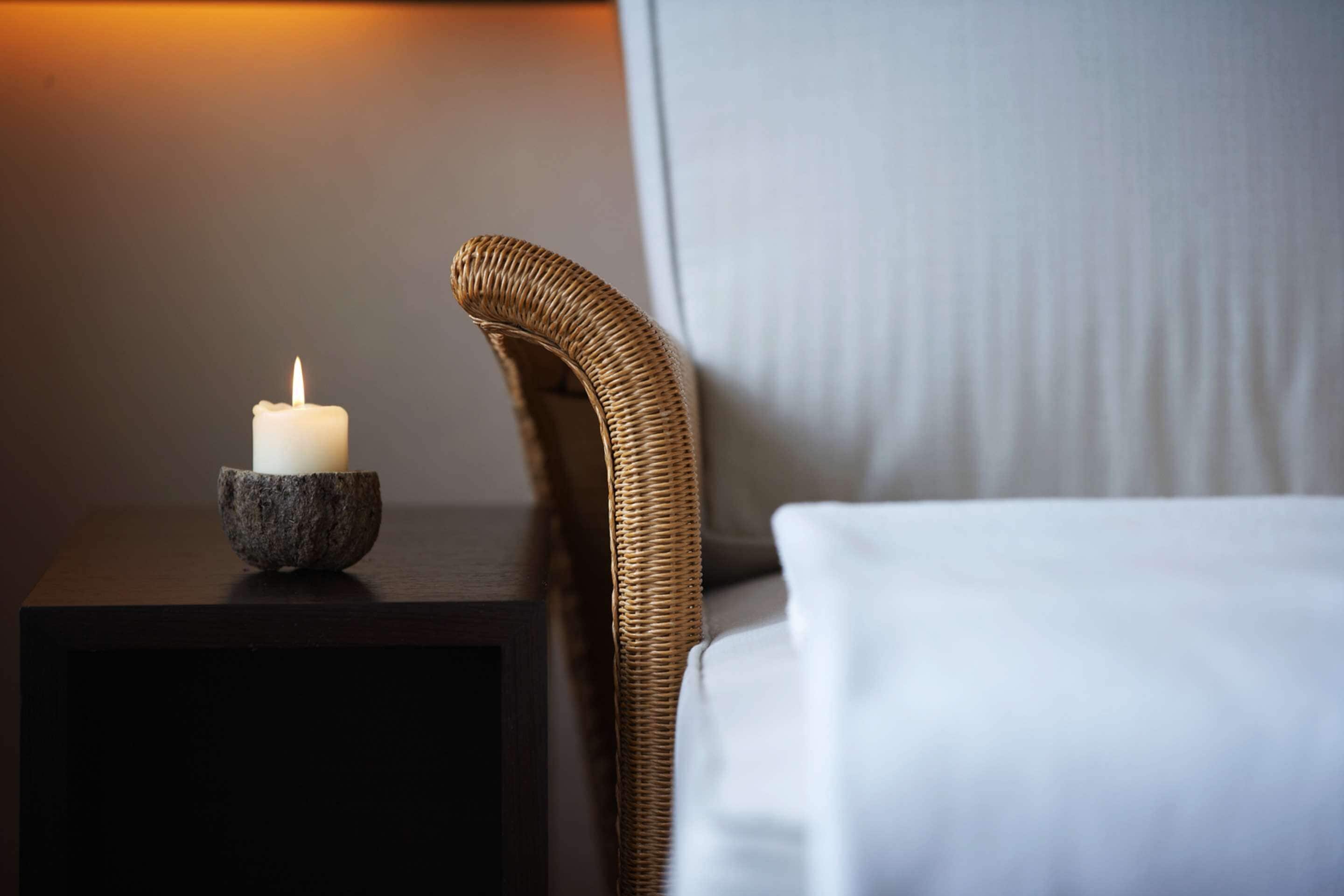 Una vela en un soporte de piedra sobre una mesita de noche de madera oscura, junto a una silla de mimbre con un cojín blanco.