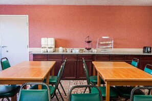 A room with a kitchen area, including a countertop with various items, a sink, and a door. There are two wooden tables with chairs, and a patterned carpet on the floor.
