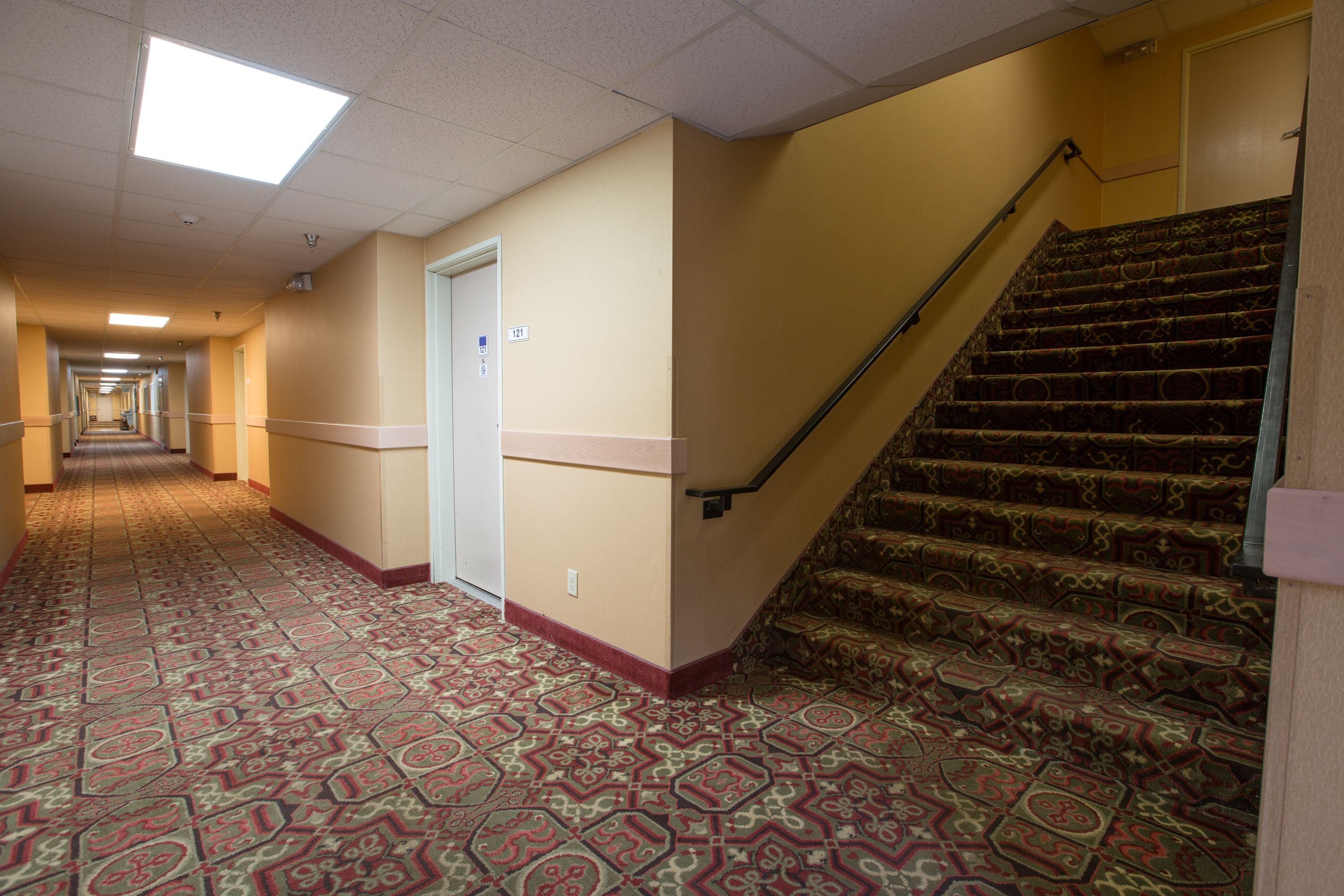 Un corridoio d'albergo con tappeto a motivi decorativi, una scala con gradini finemente lavorati e una porta con il numero 121.