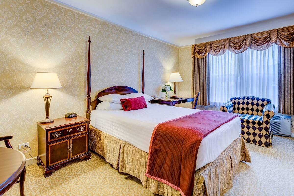 A hotel room with a four-poster bed, a nightstand with a lamp, a chair, and a large window with curtains.