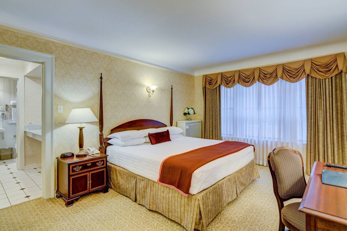 A hotel room with a large bed, a desk, a chair, a nightstand, a lamp, and a mirror.
