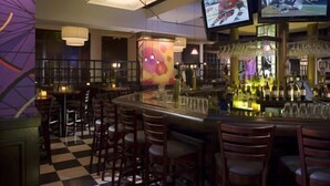 A bar area with a checkered floor, wooden chairs, a bar counter, and two televisions mounted on the wall.