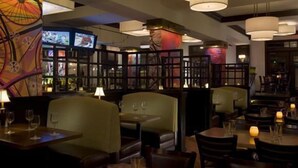 A dimly lit restaurant with booths and tables, featuring a large clock and a television screen on the wall.