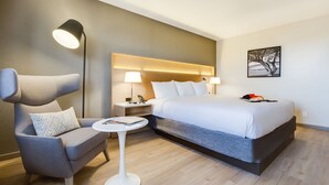 Una habitación de hotel moderna con una cama grande, un sillón gris, una mesita redonda y una lámpara de pie.