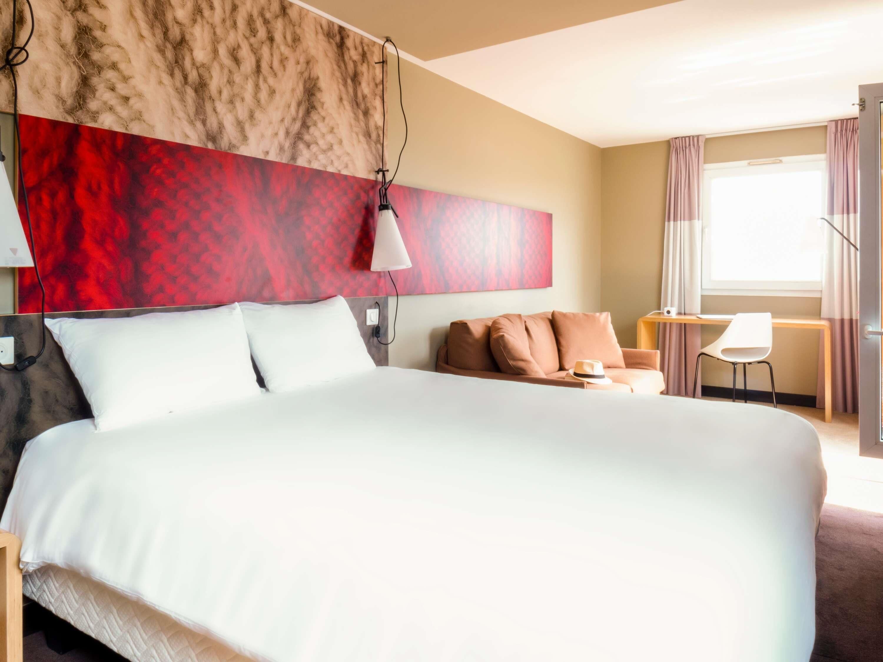 Habitación de hotel con una cama grande, un escritorio con una silla, una pared de color rojo y beige, y una ventana con cortinas.